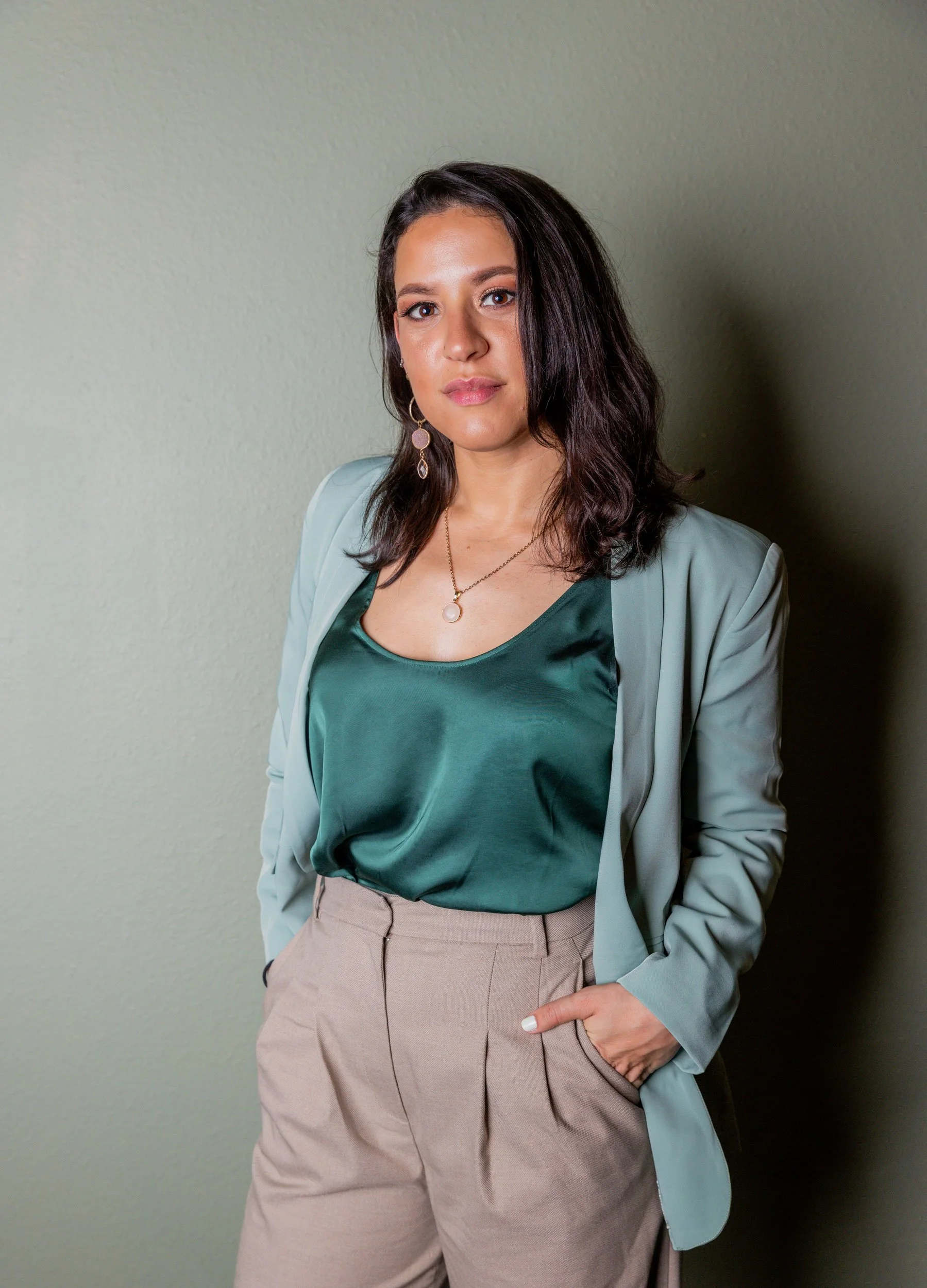A woman with shoulder-length dark brown hair, wearing a satin emerald green top, beige high-waisted trousers, and a light blue blazer, standing against a light green wall.