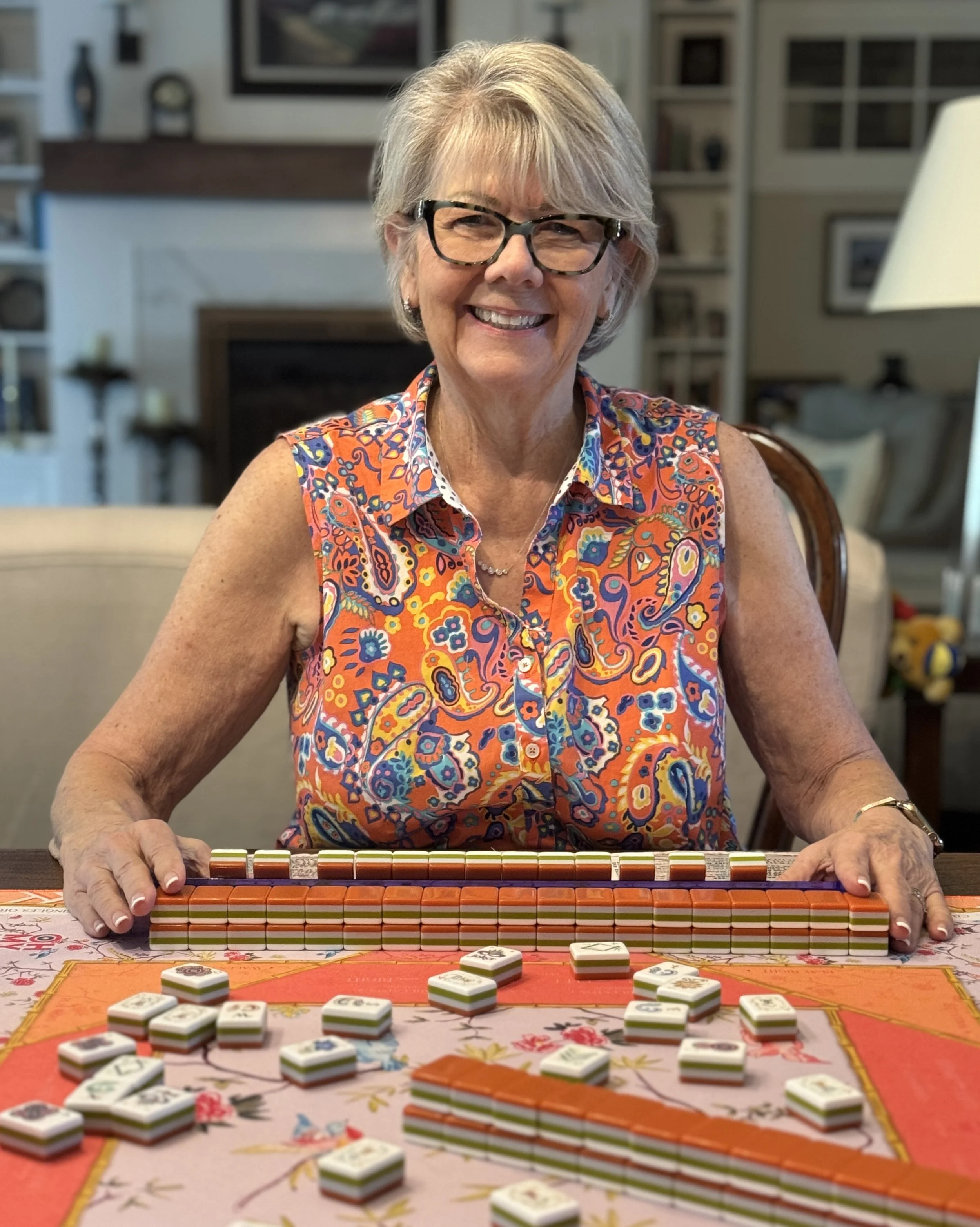 Smiling woman sitting at a table with Mahjong tiles in front of her, ready to play the game.