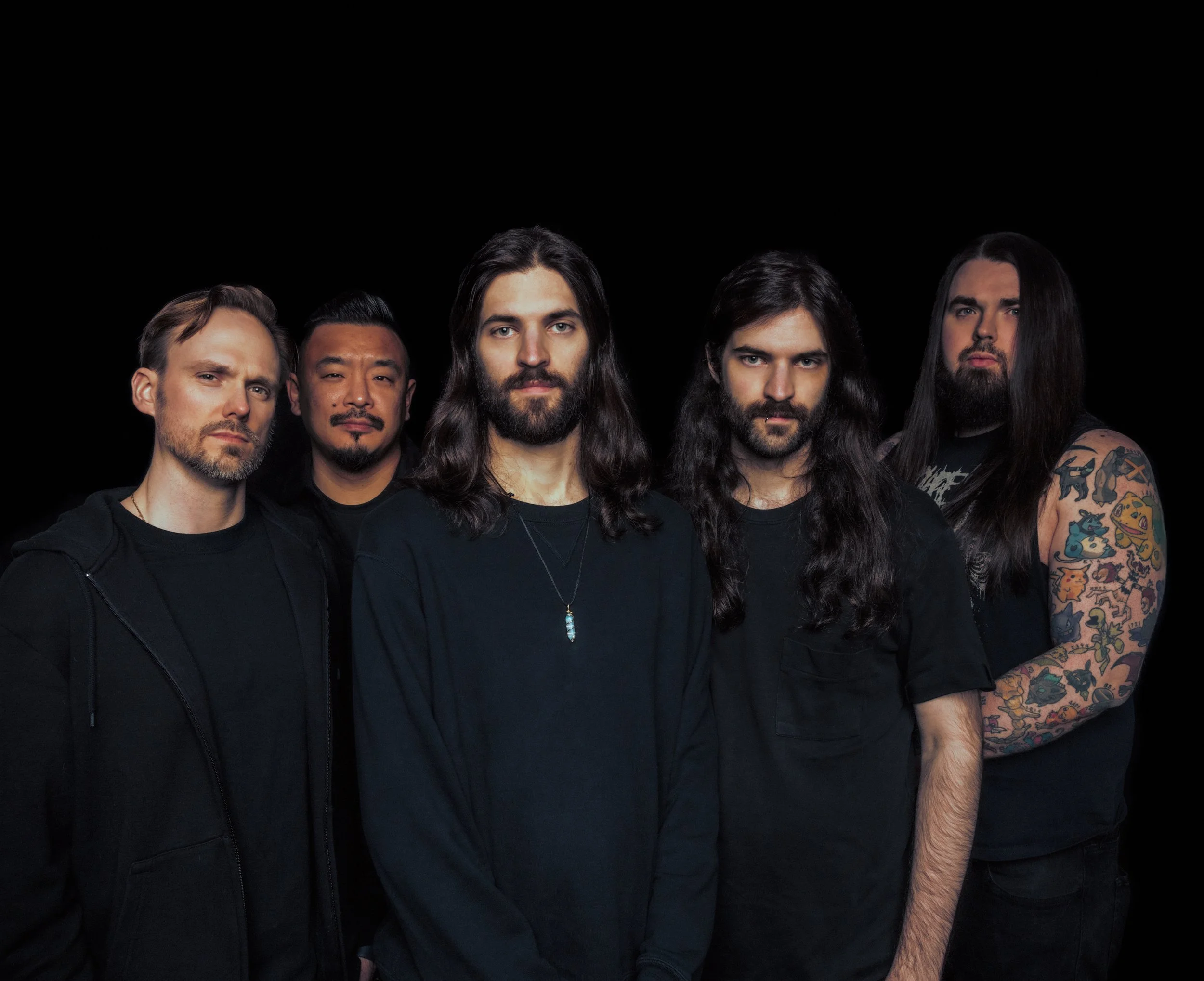 Five men with long hair and beards, dressed in black, standing against a black background, posing for a group photo.