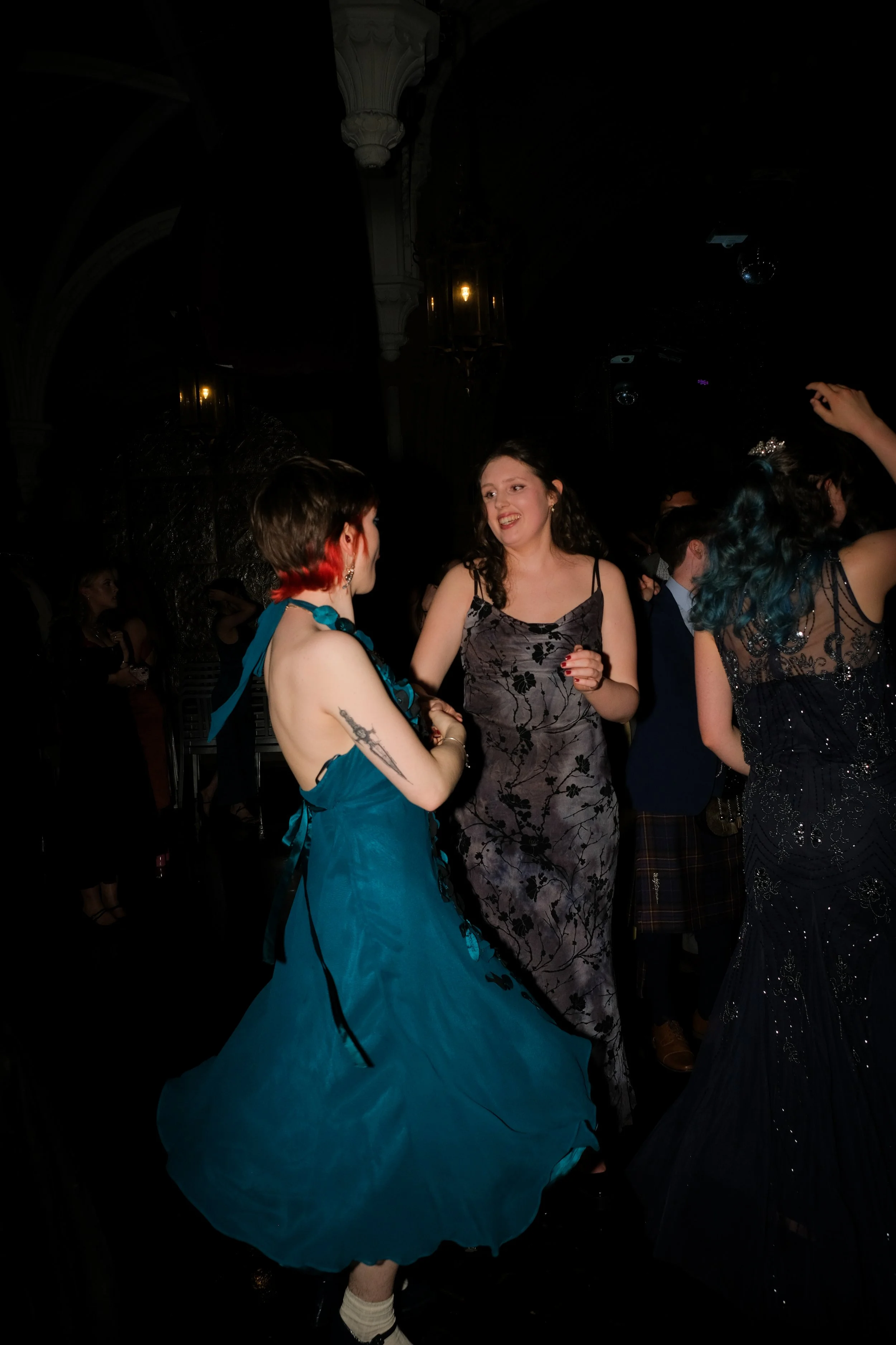 Two women in colorful dresses dancing and talking at a dark, elegant party or event.