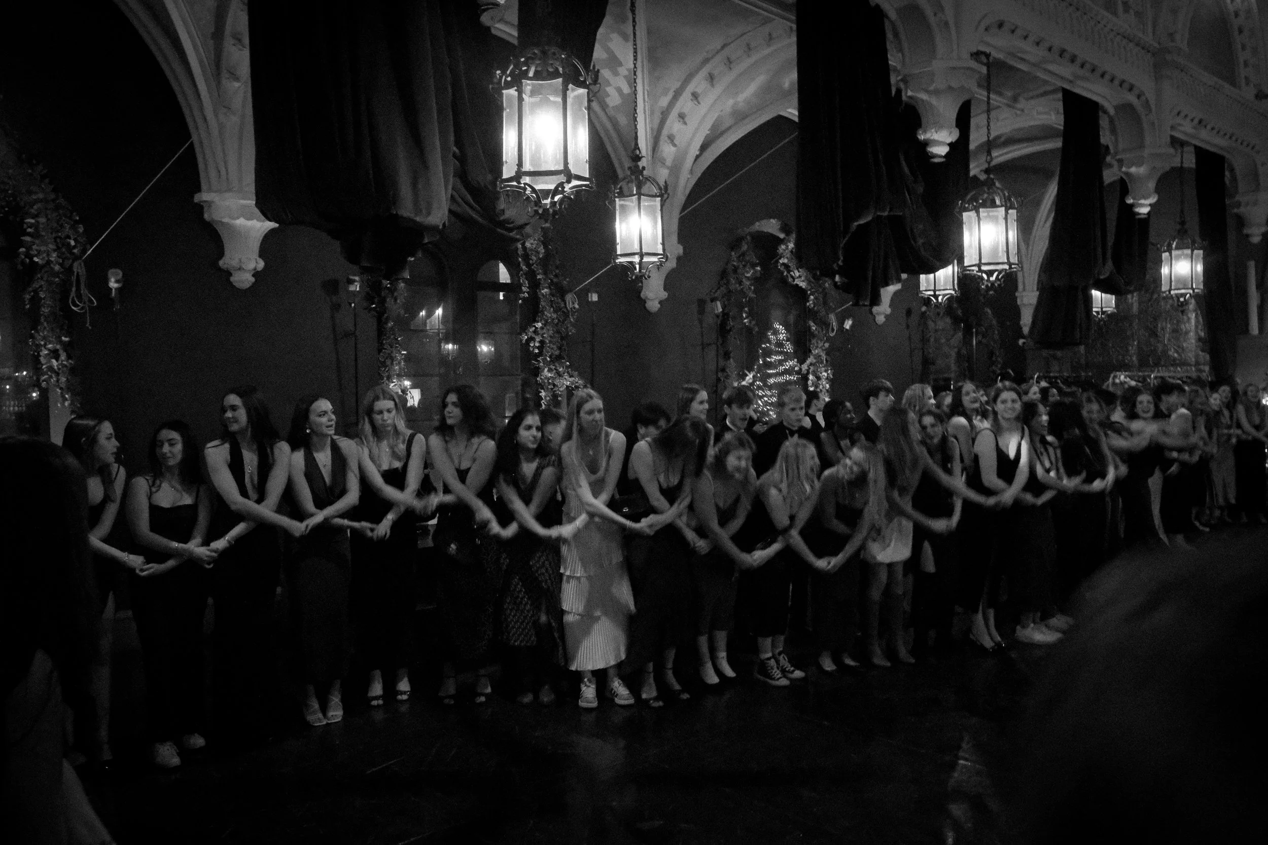 A group of young women standing in a line, holding hands, in an ornate, dimly lit room decorated for a holiday event with Christmas trees and hanging lanterns.