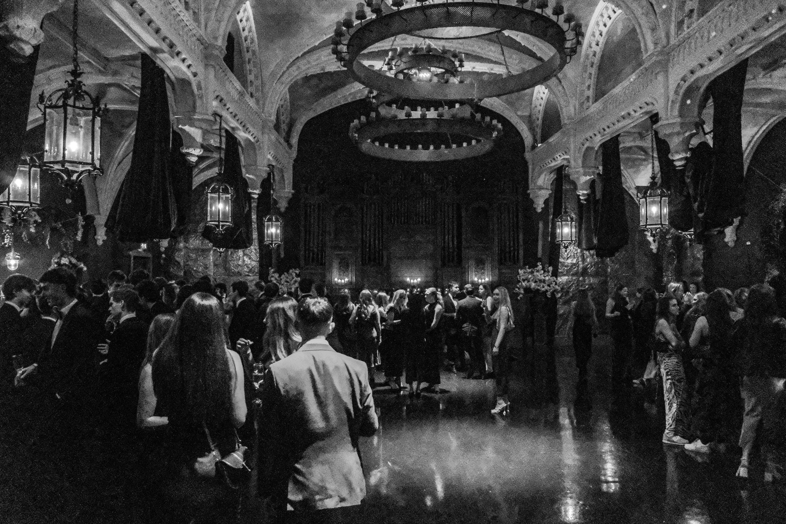Black-and-white photo of a crowded indoor social gathering or party with people dancing and mingling in a grand, ornate hall decorated with chandeliers, hanging lanterns, and floral arrangements.