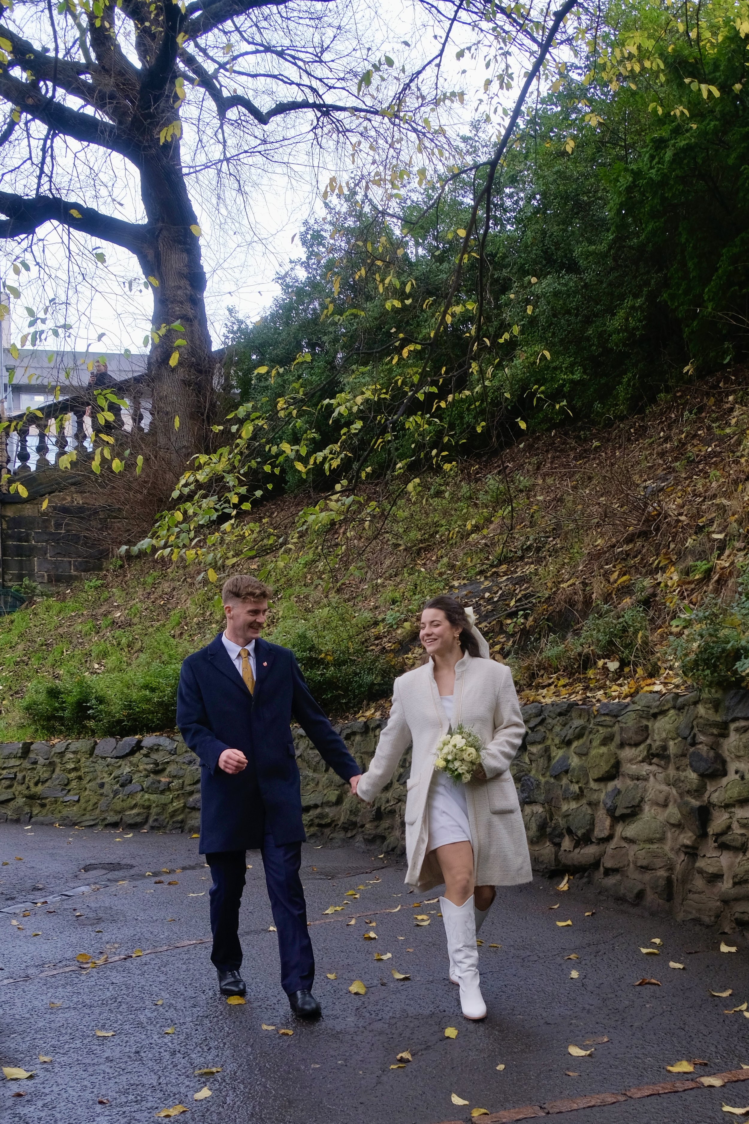 A bride and groom walking hand in hand outdoors, smiling and dressed in wedding attire, with a large tree and a stone wall in the background on an overcast day.