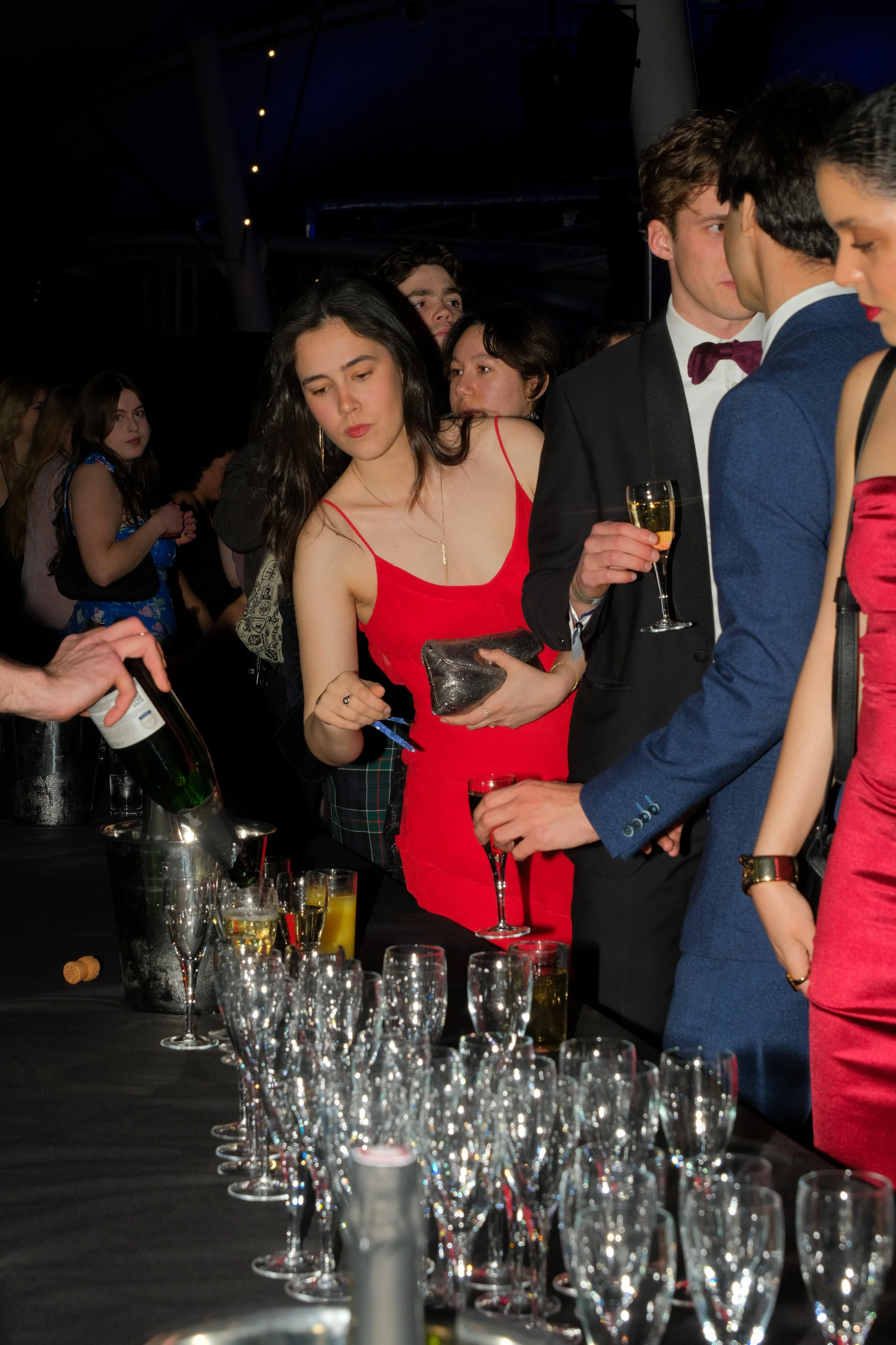 People dressed formally at a party, waiting to be served champagne and drinks from a table with champagne glasses, with a dark background and string lights overhead.