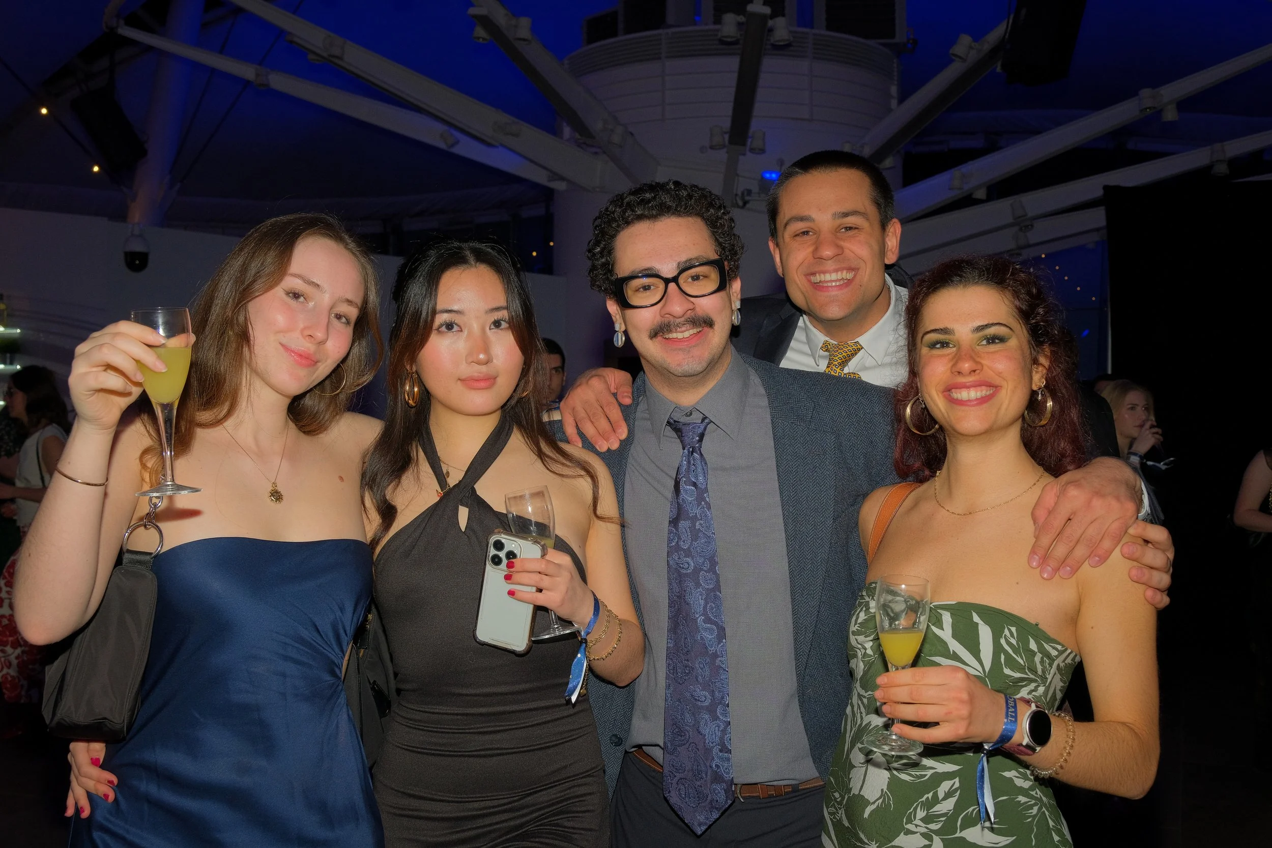 Group of five diverse people at a party, smiling and posing for a photo, holding drinks, in a dimly lit indoor setting.