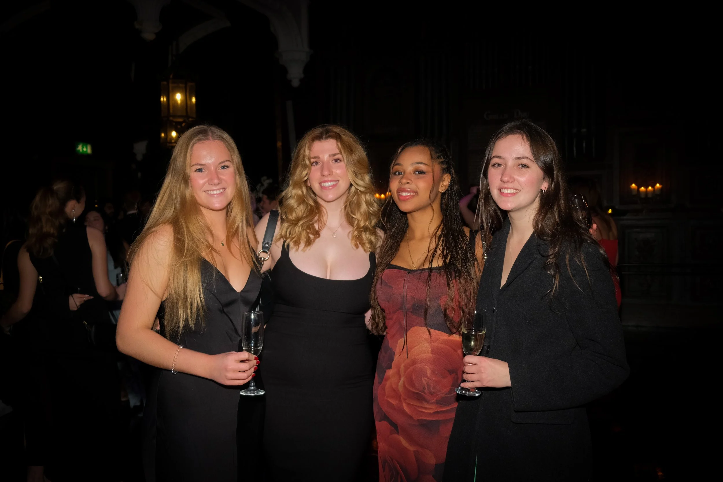 Four women at a party holding champagne glasses, standing close together in a dimly lit room with candles in the background.