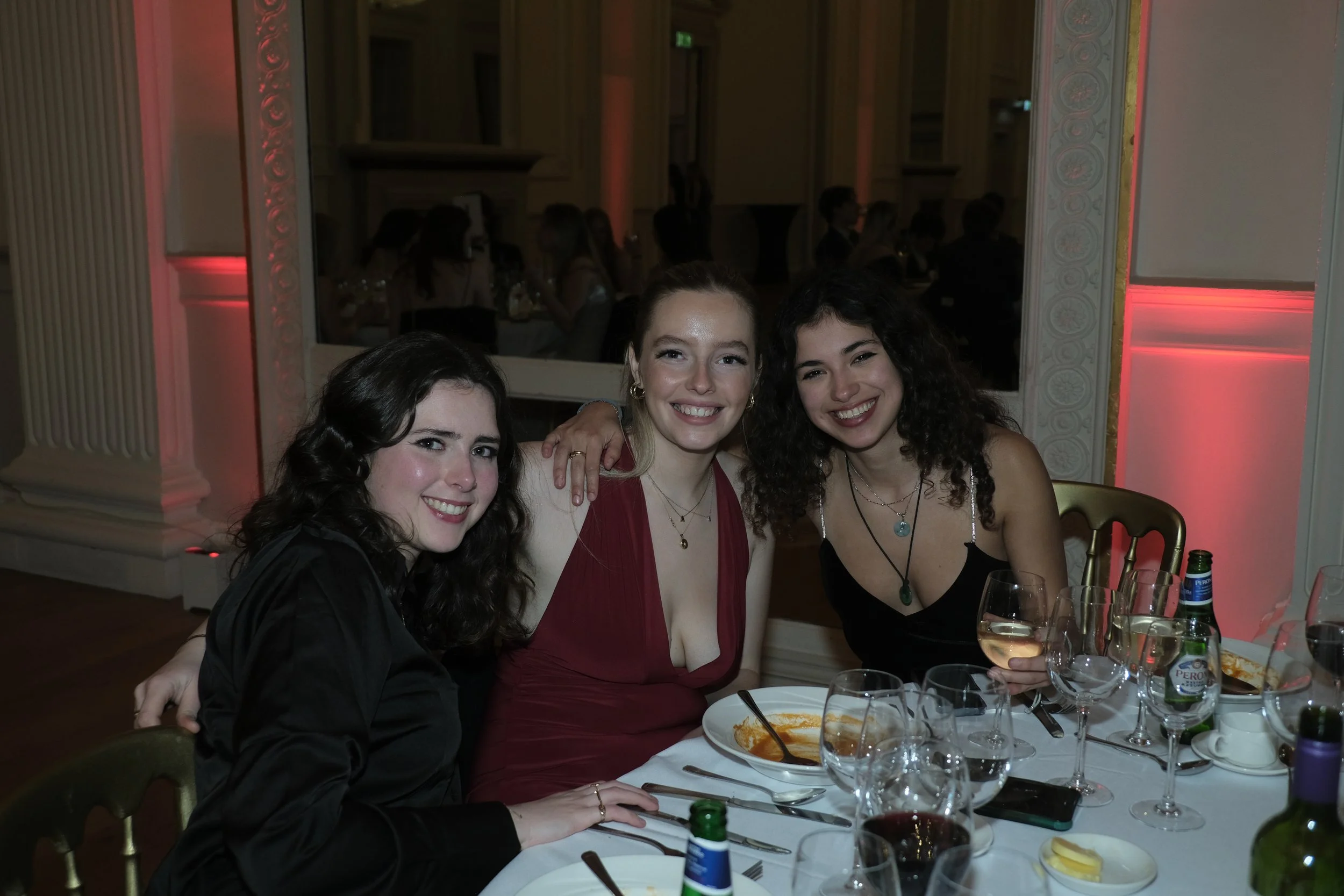 Three women smiling at a dinner table with food and drinks at an elegant event.