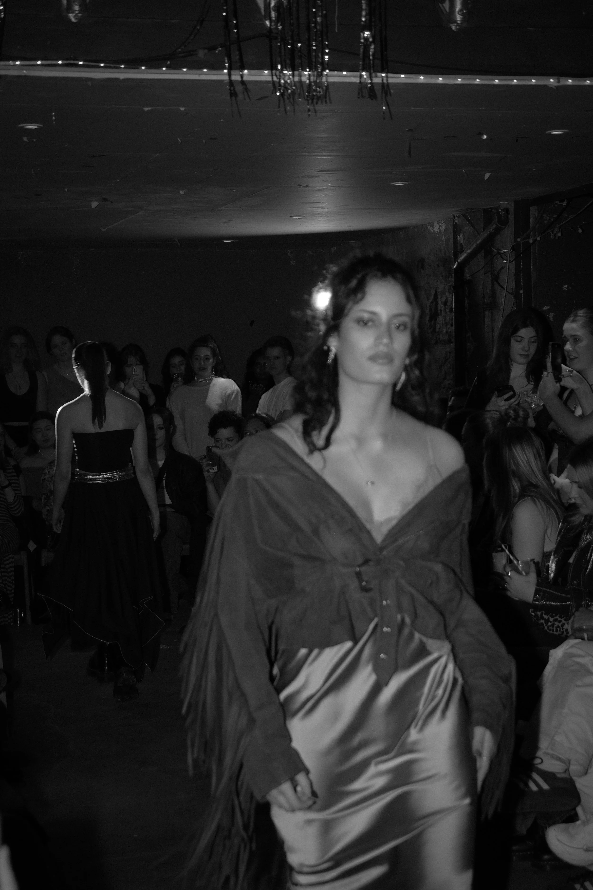 A woman walking on a runway during a fashion show, with an audience watching and some people taking photos in the background.