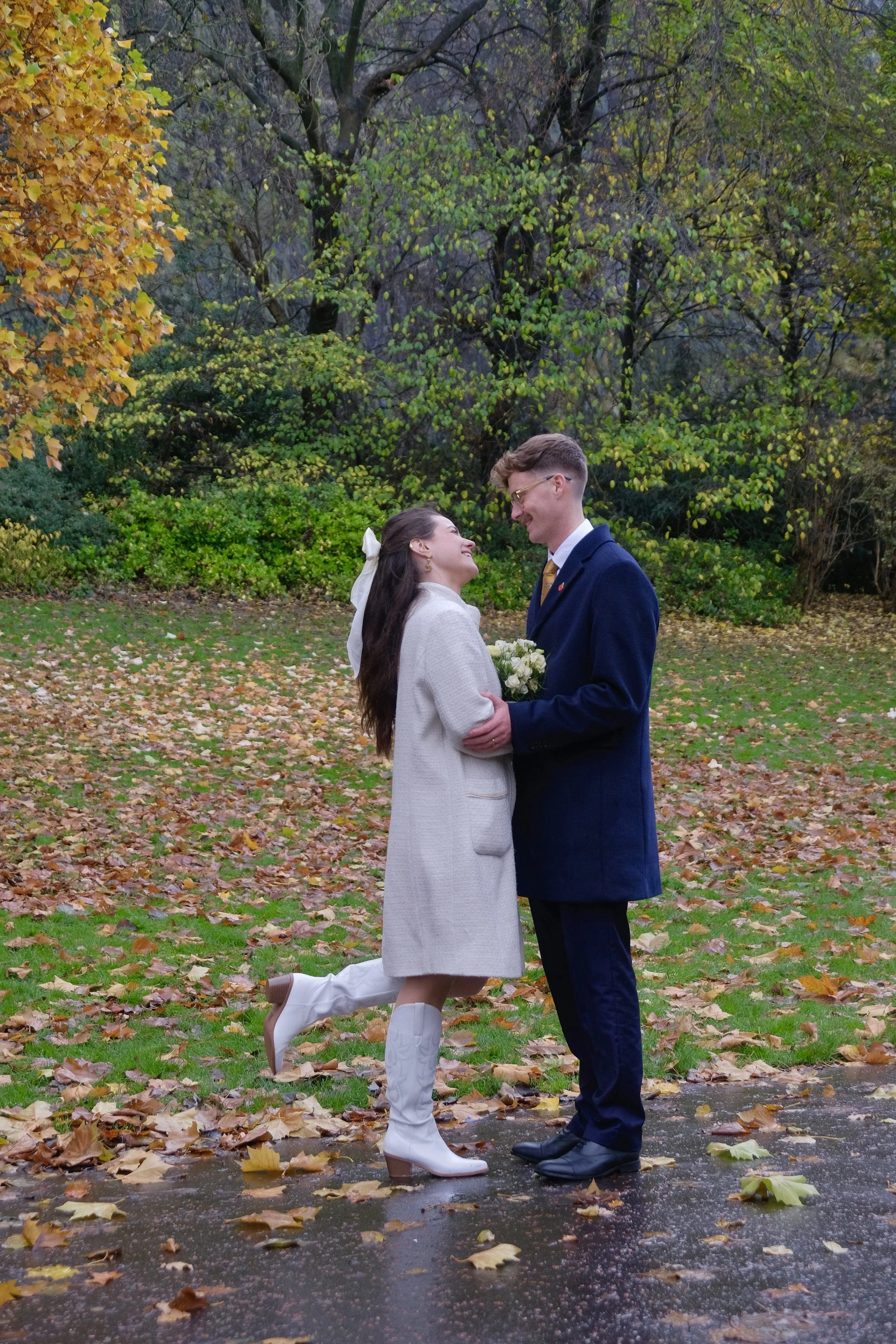A couple dressed in vintage attire sharing a romantic moment outdoors in an autumn park, with colorful fall leaves on the ground and trees in the background.