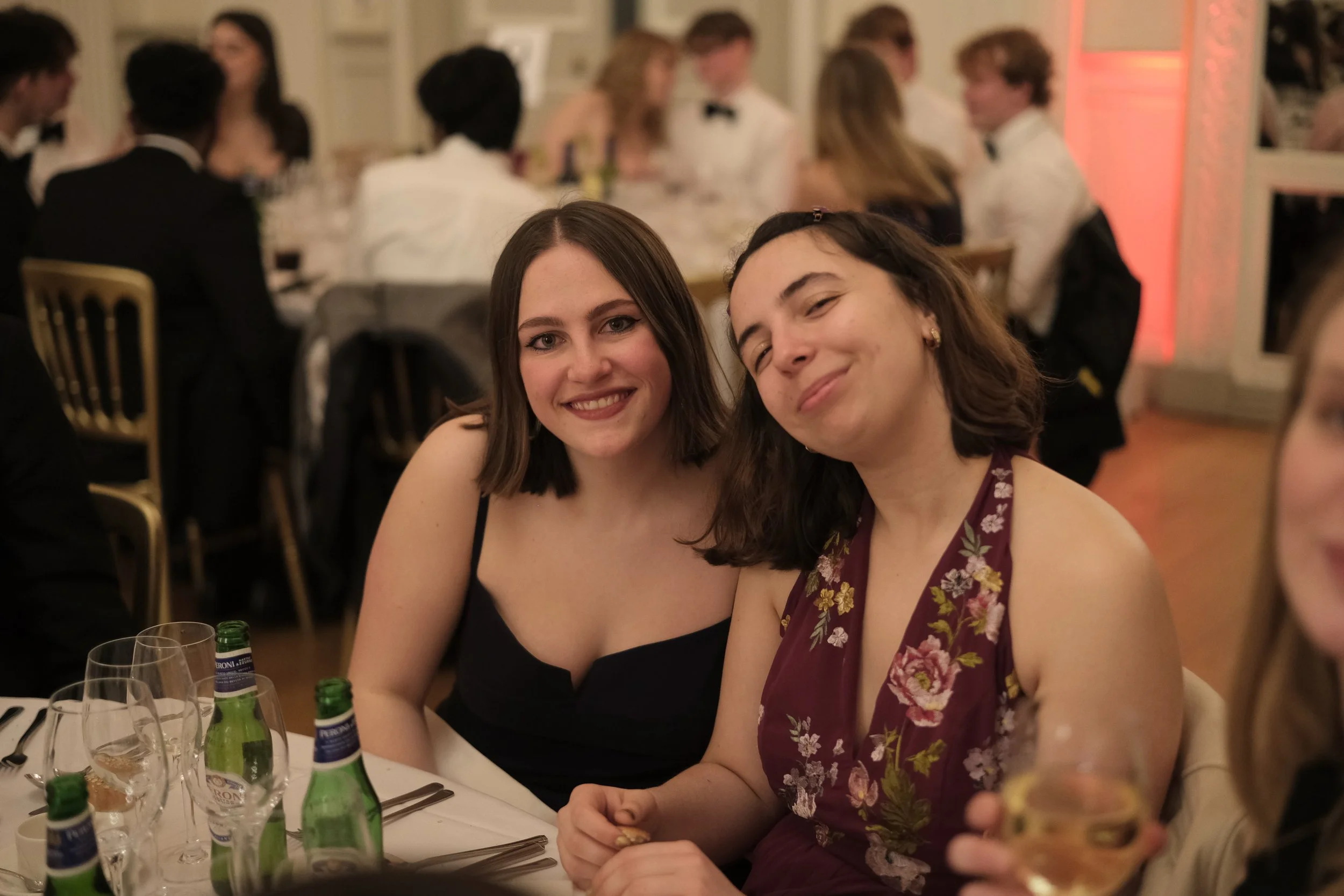 Two women sitting at a dinner table, smiling at the camera, with a lively gathering of people in formal attire in the background.
