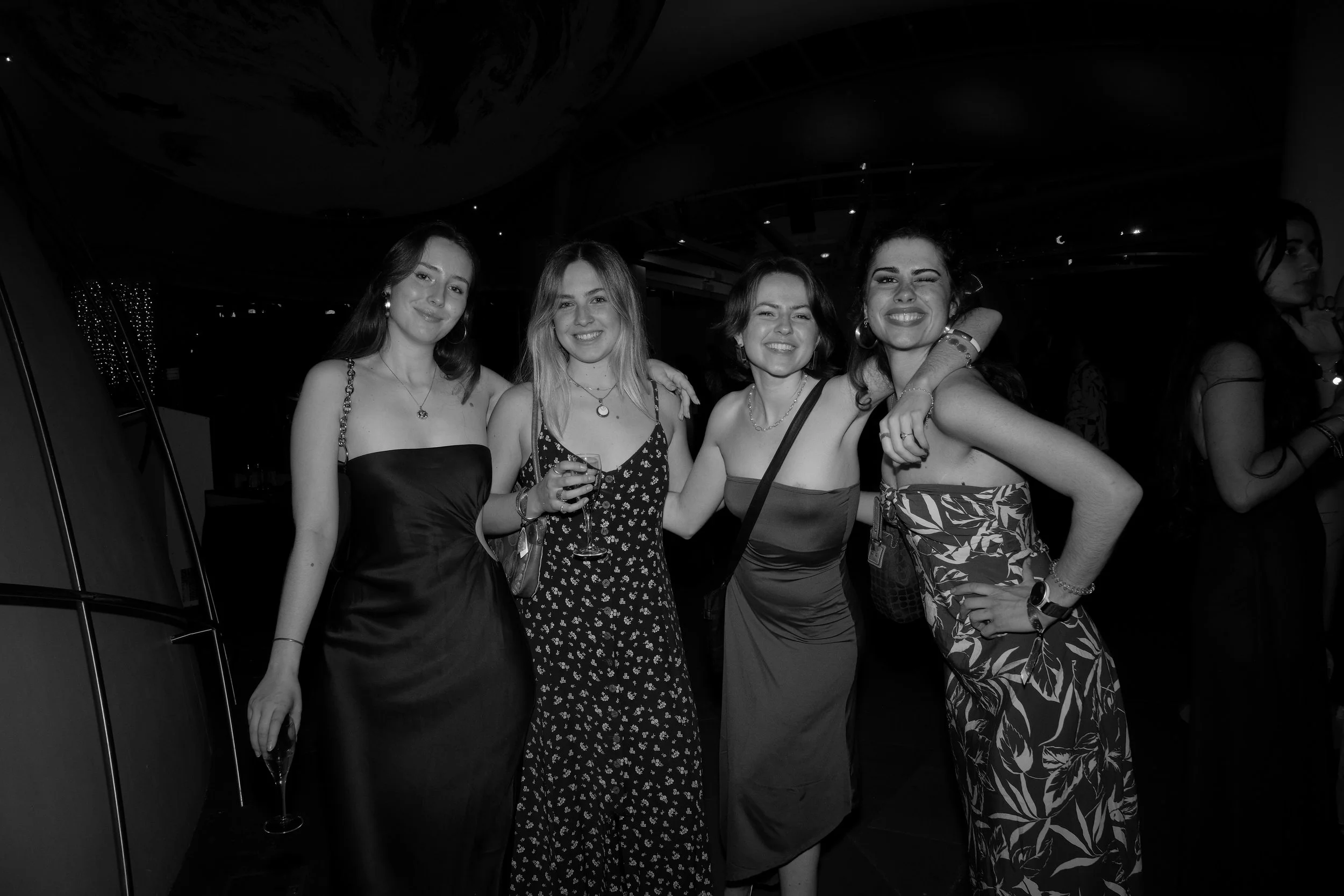 Four women in dresses smiling and posing together at a social event, with one holding a glass of wine.