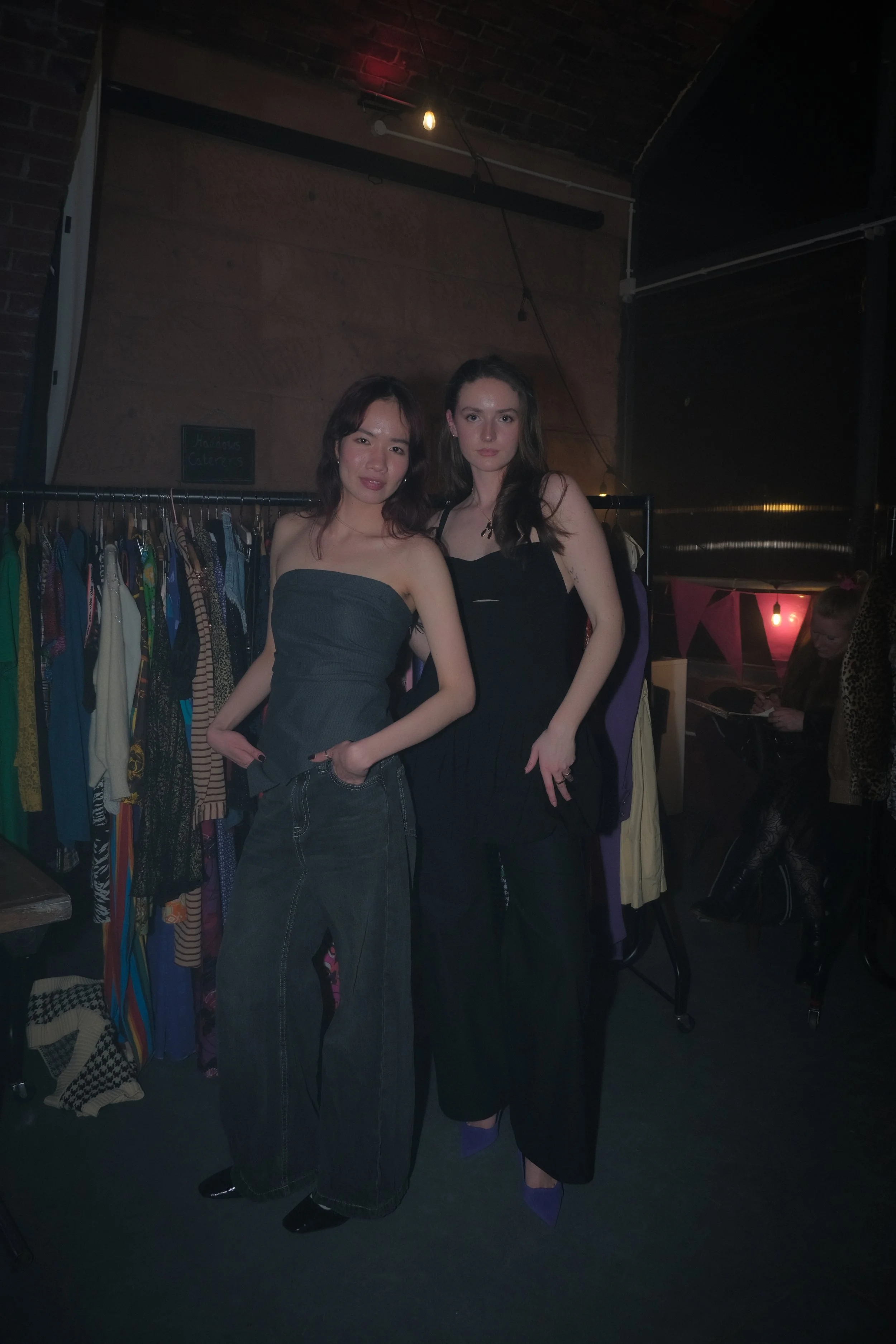 Two women standing in front of a clothing rack with colorful clothes, inside a dimly lit room.