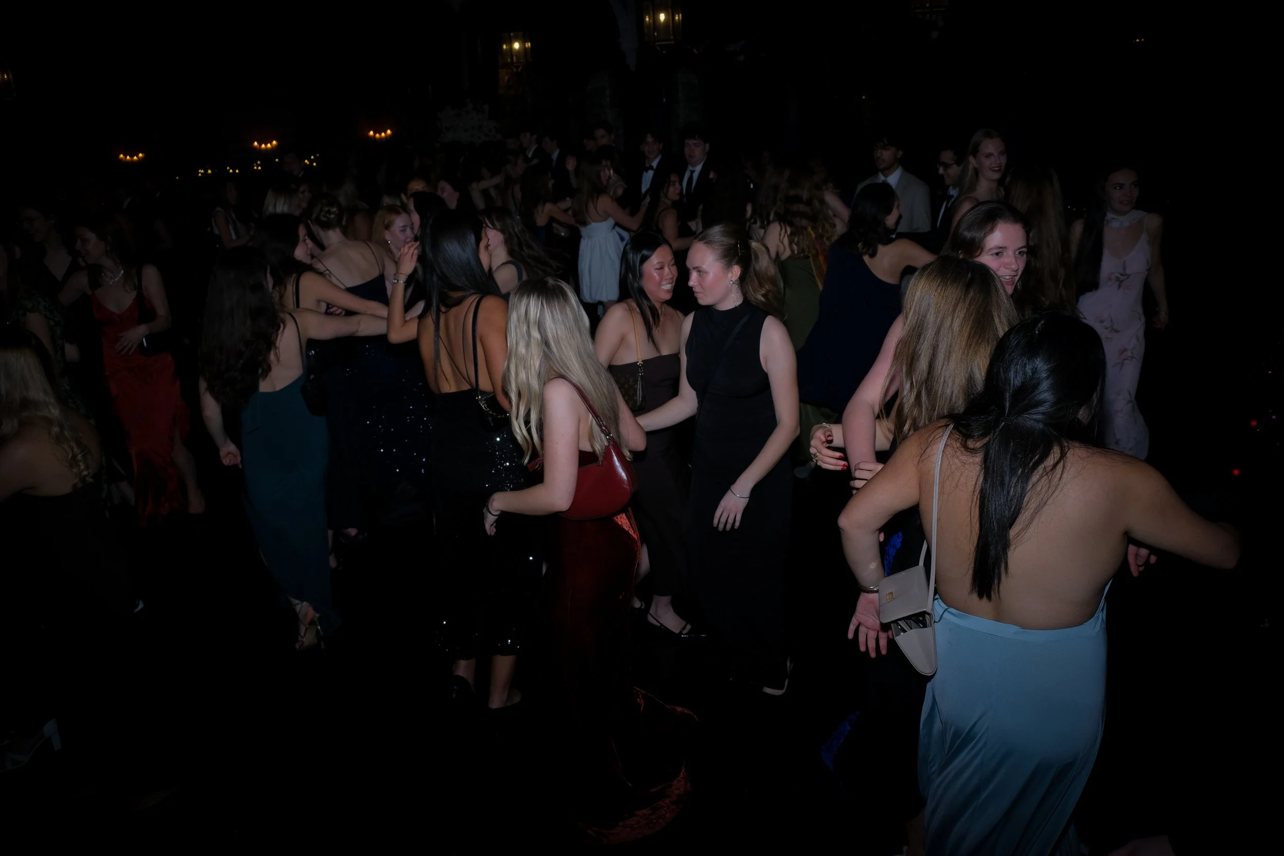 A group of young women dancing and socializing at a dark, elegant party or event, mostly dressed in formal and semi-formal attire.
