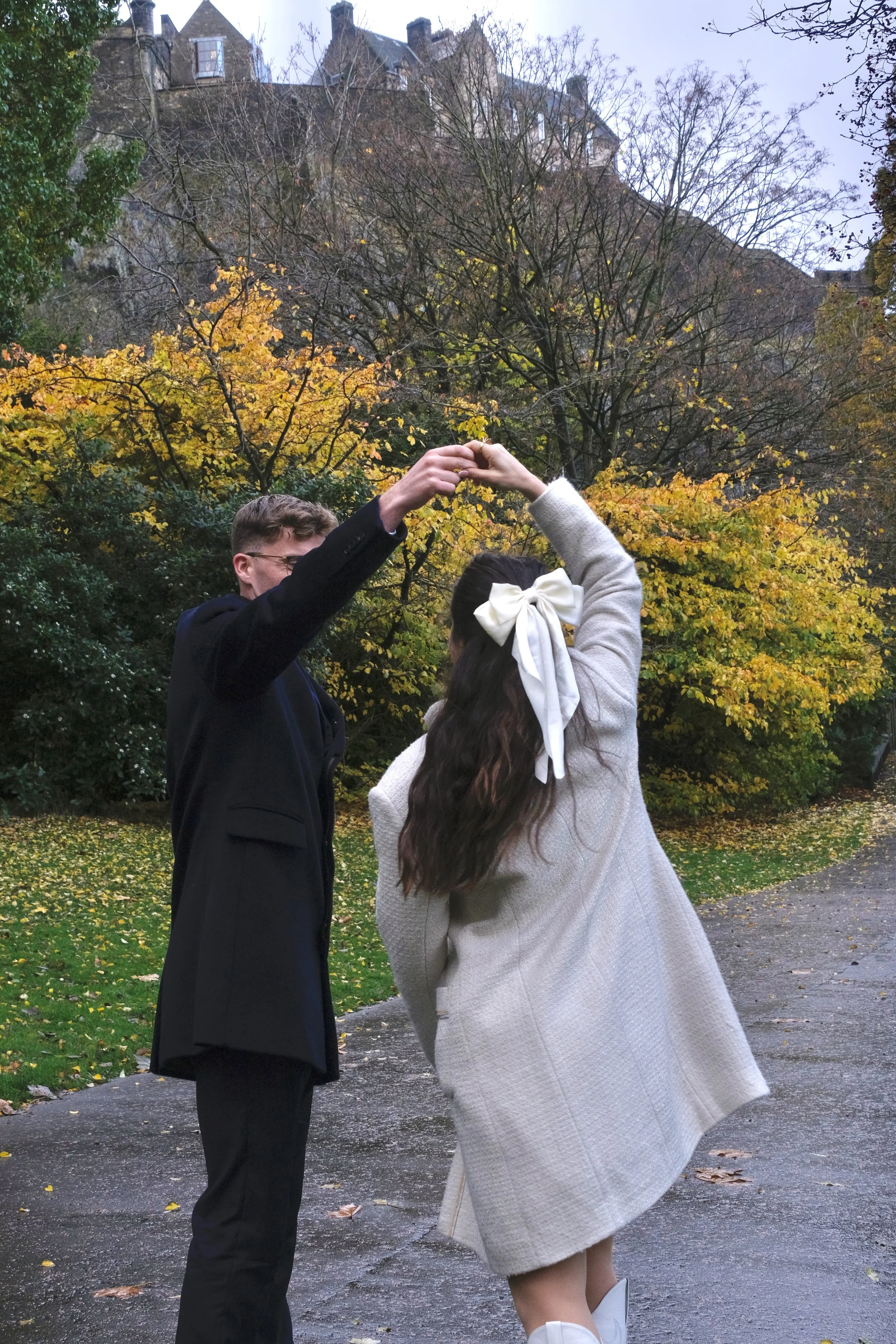 A young man and woman are dancing outdoors on a paved path, surrounded by fall-colored trees and a building on a hill in the background. The woman has long brown hair tied with a large white bow and is wearing a light-colored coat. The man has short