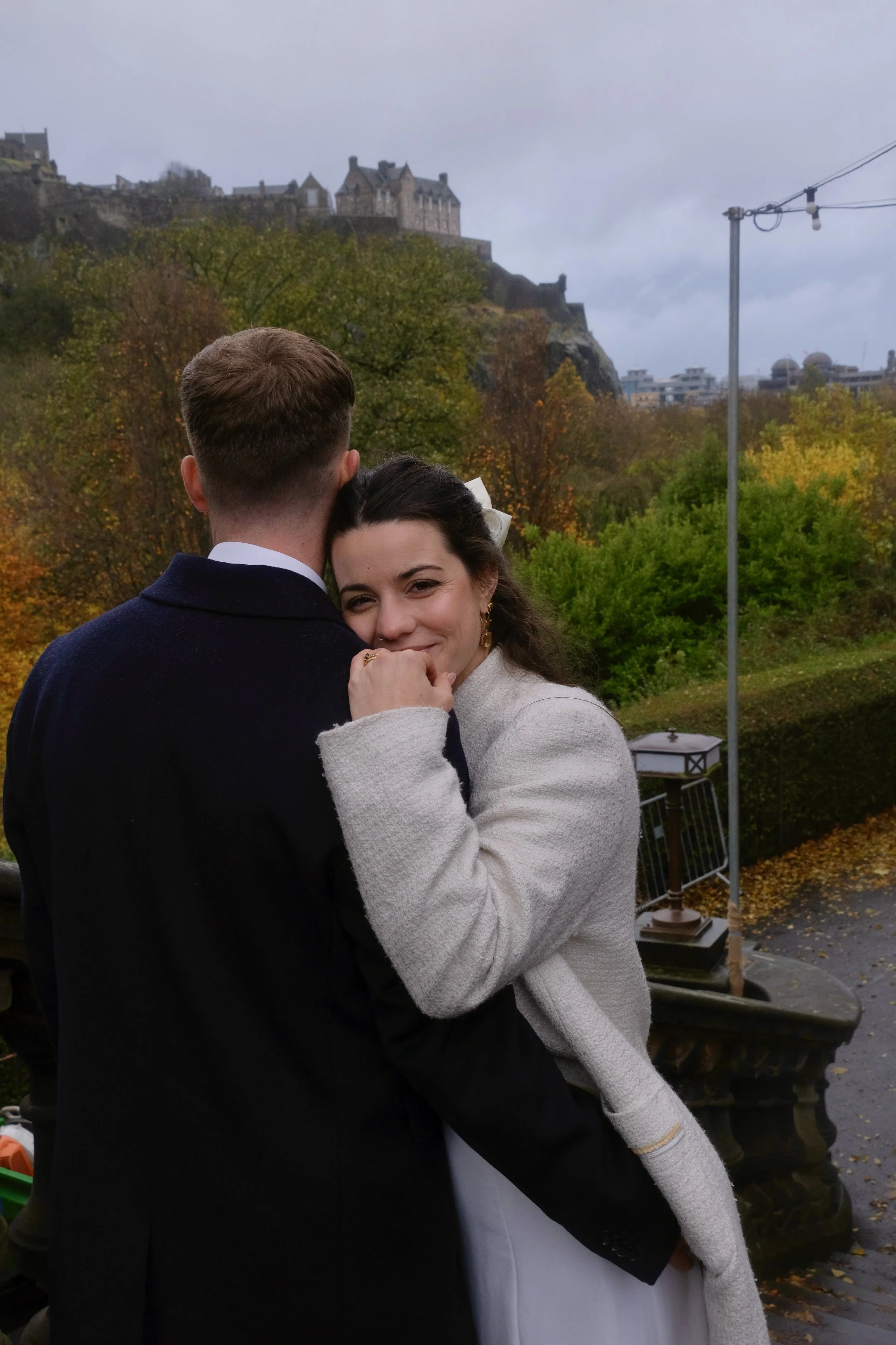 A woman smiling and hugging a man from behind outdoors, with a scenic background of trees with autumn foliage and a castle on a hill.