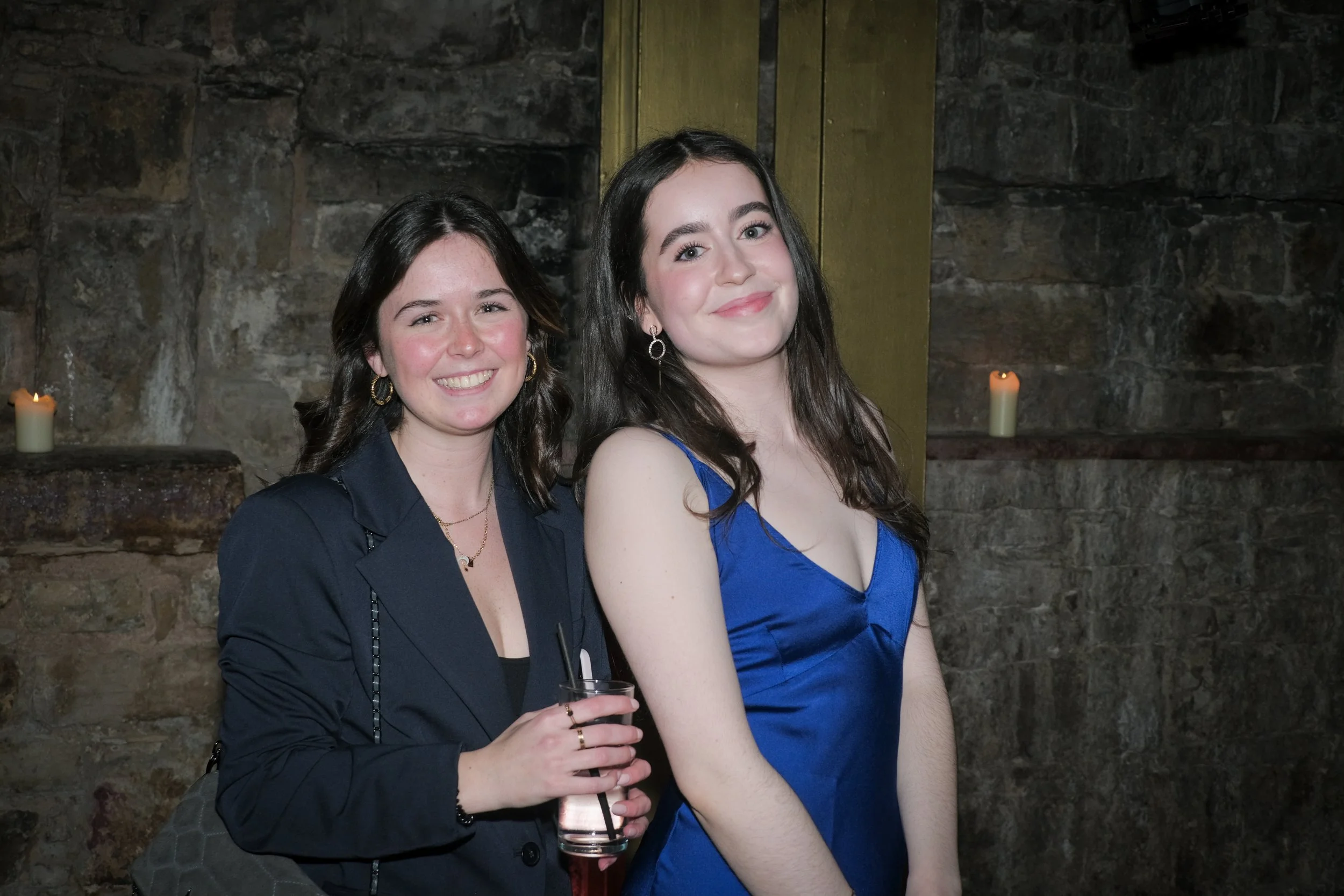 Two young women smiling at a social event, standing against a stone wall with lit candles in the background.