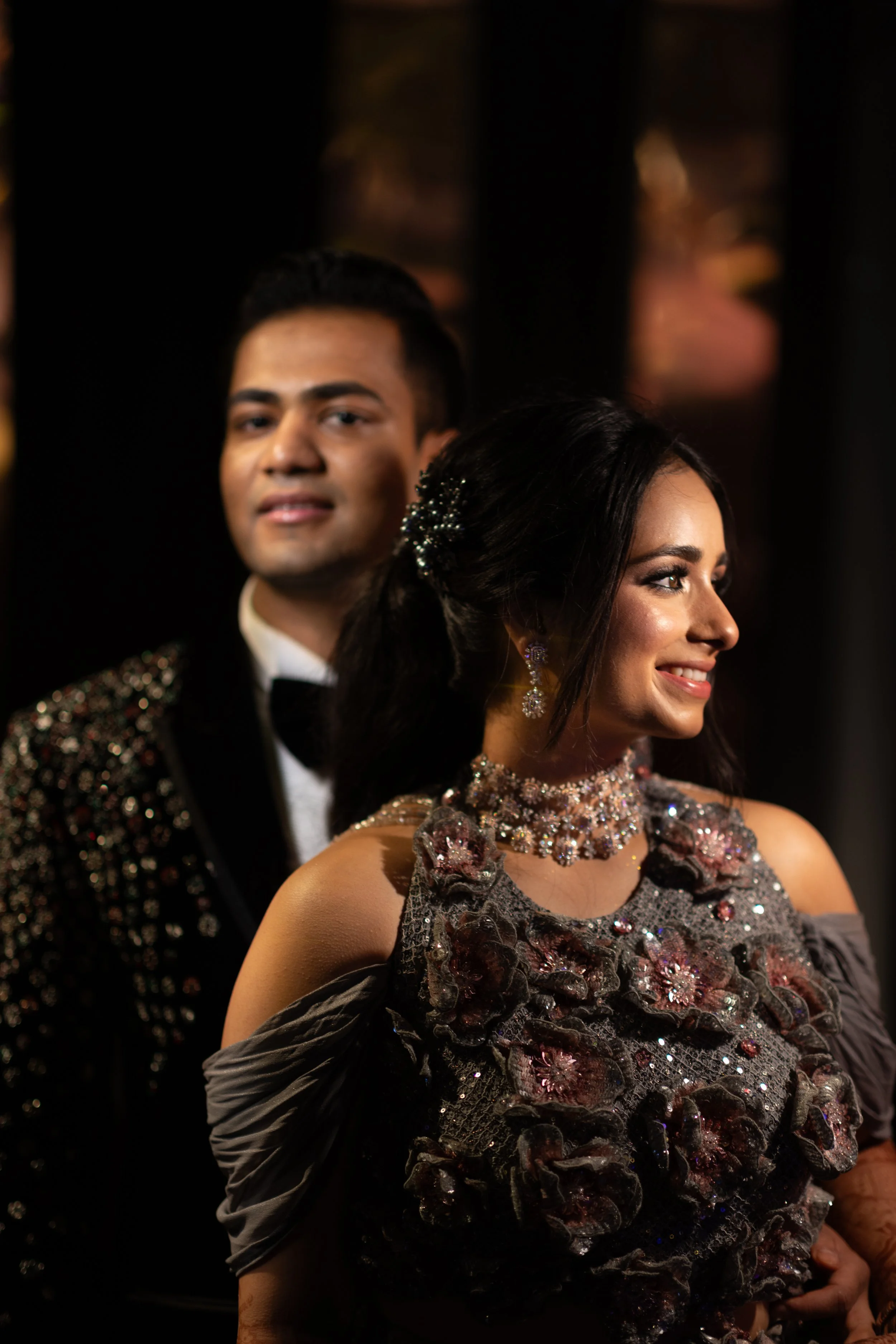 A woman dressed in an ornate, floral, embroidered gown and jewelry, smiling, looking to the side. Behind her, a man in a tuxedo with a sparkling jacket, also smiling, slightly out of focus.
