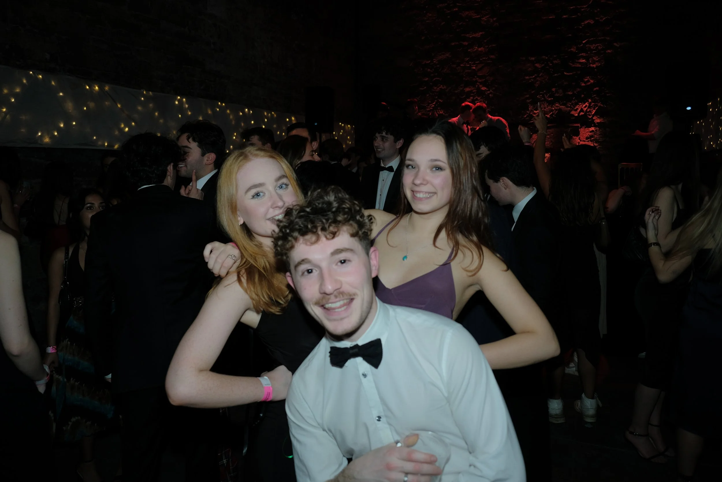 Young man in white shirt and black bowtie smiling at camera, flanked by two young women at a formal event with a crowd of people dancing and socializing in the background.