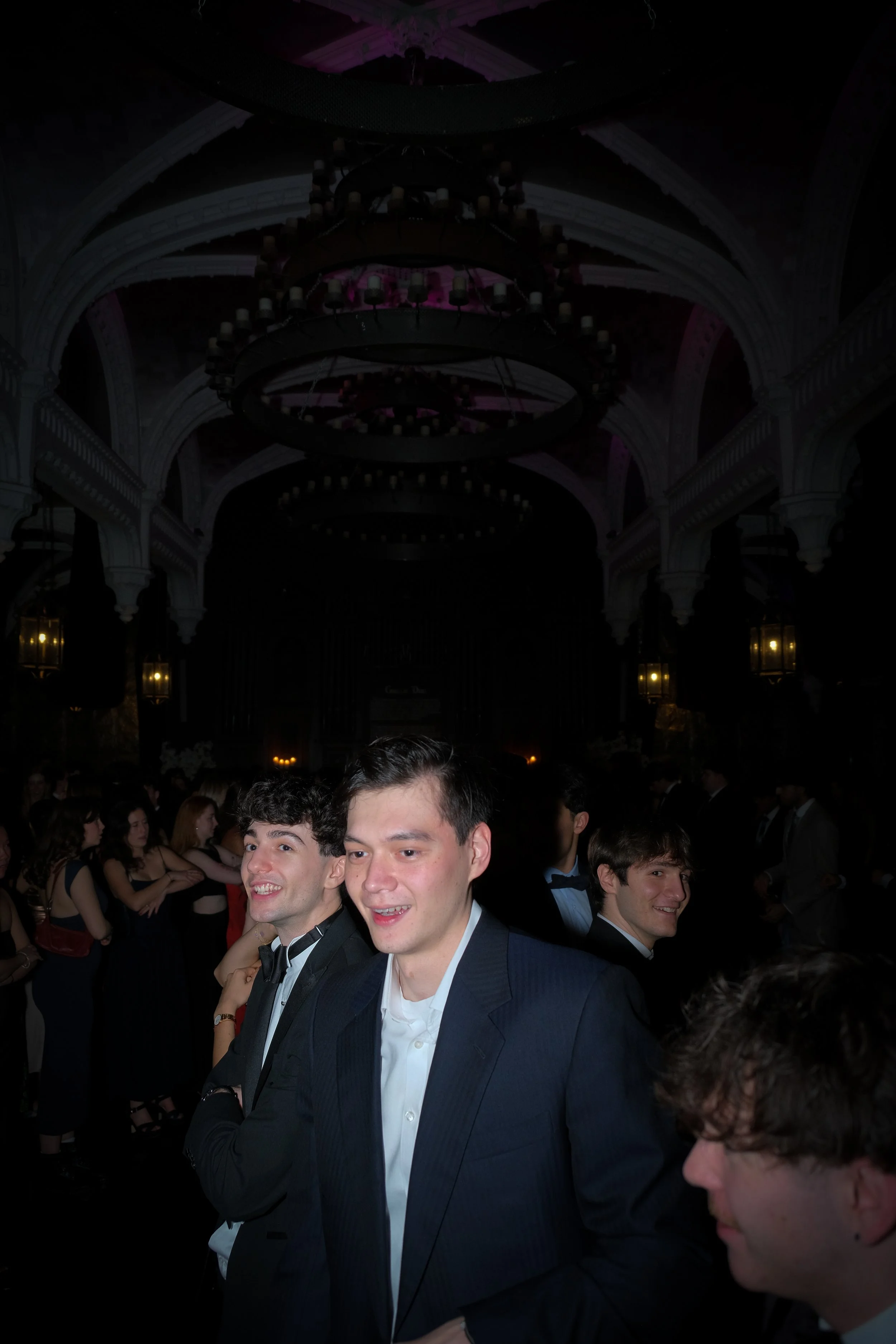 Young men in suits at a formal dance or prom, standing in a large hall with chandeliers and decorated architecture.