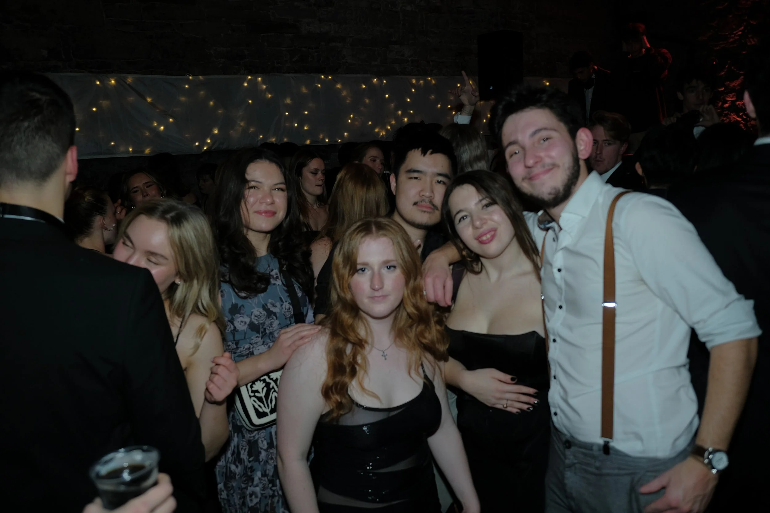 Group of young adults enjoying a party or celebration indoors at night with string lights in the background.