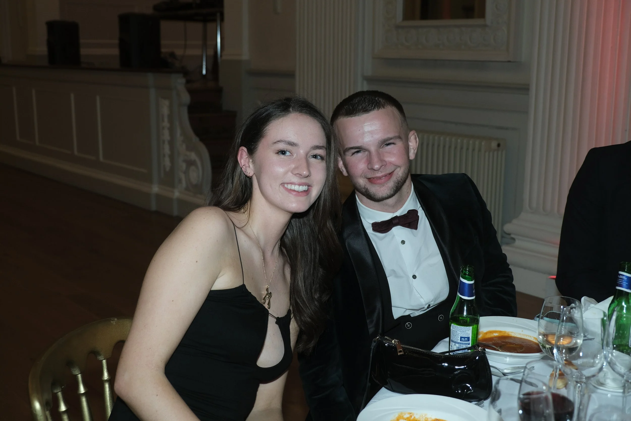 A young woman and man smiling at a formal event, sitting at a table with food and drinks, dressed in elegant attire.