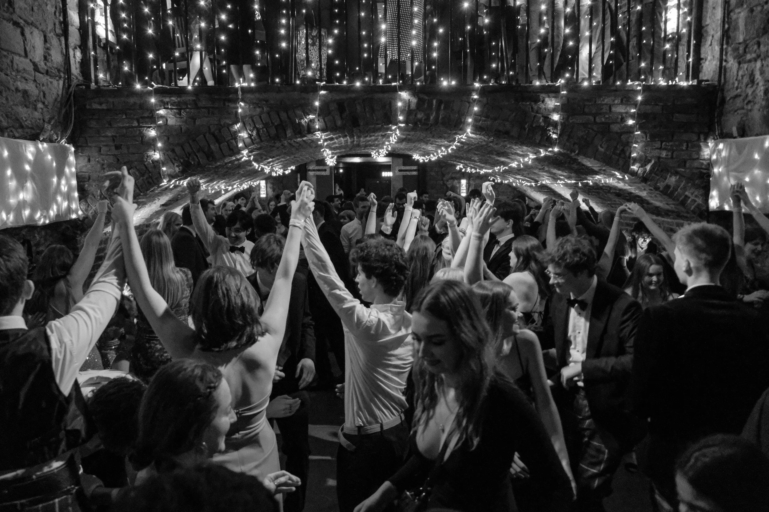 People dancing at a celebration or party in a rustic, brick-walled venue decorated with string lights.