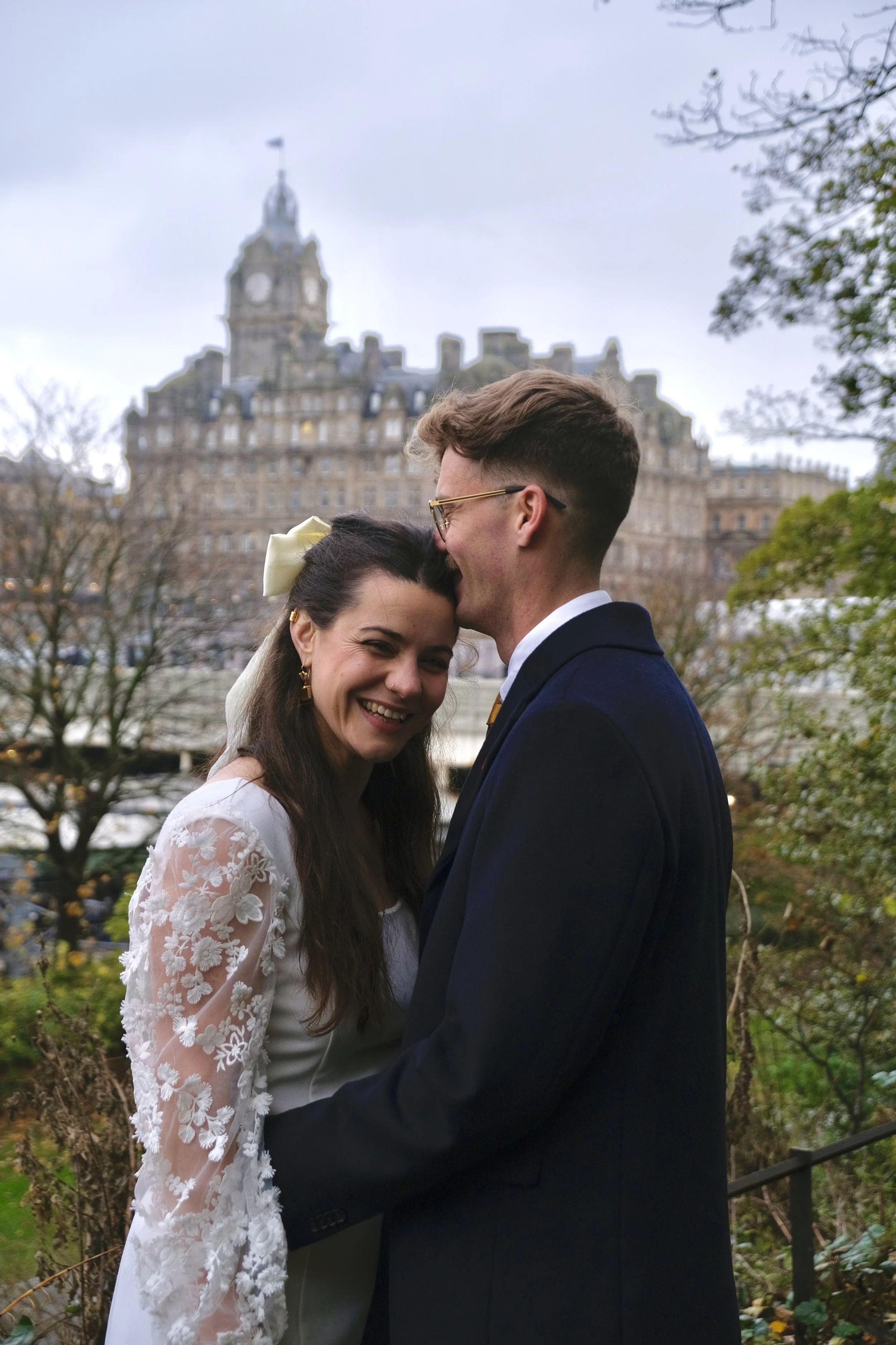 A happy couple on their wedding day, with the bride in a white lace dress and the groom in a dark suit, embracing outdoors with a historic building in the background.
