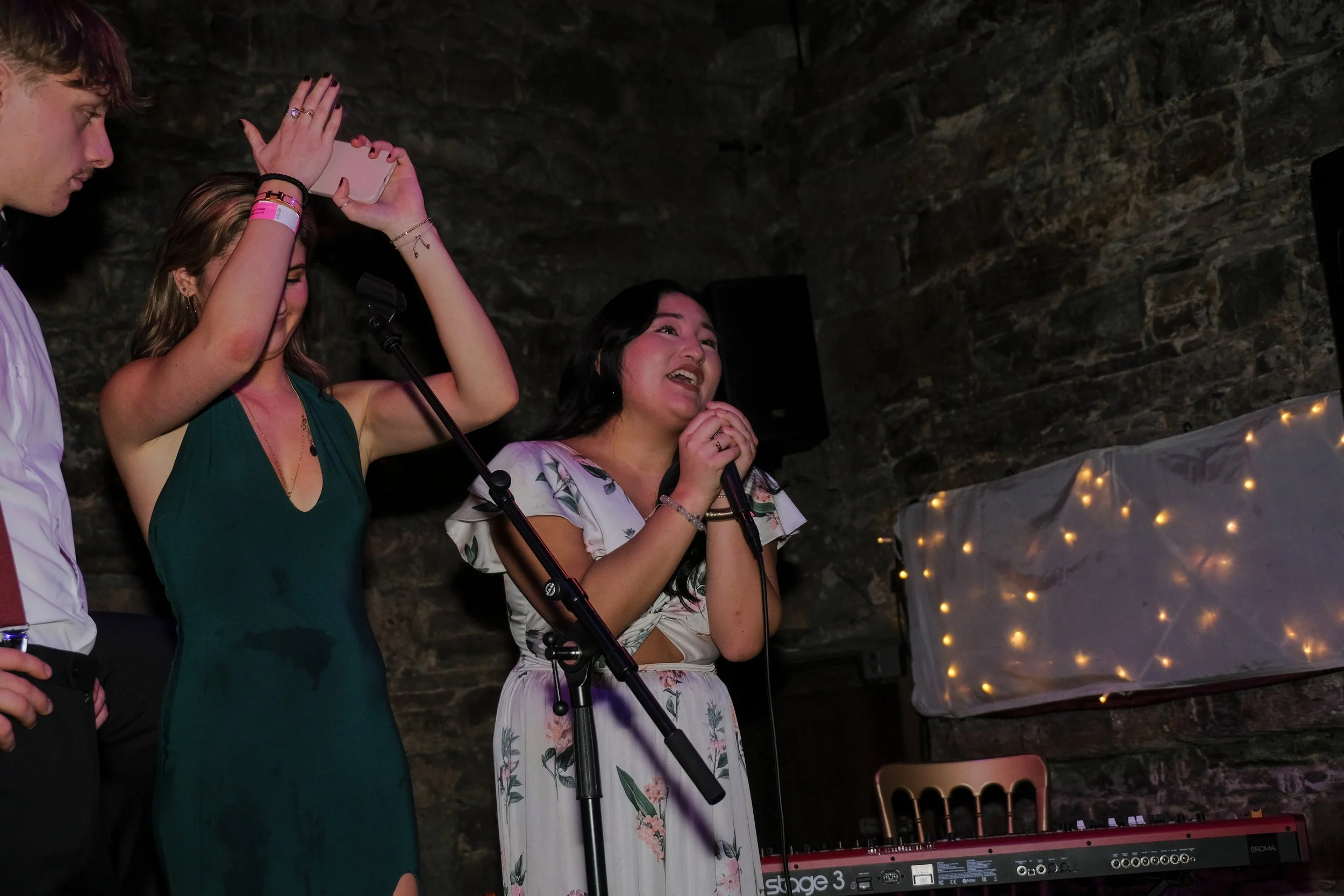 Women singing into microphones at a party or event with a keyboard and string lights in the background.