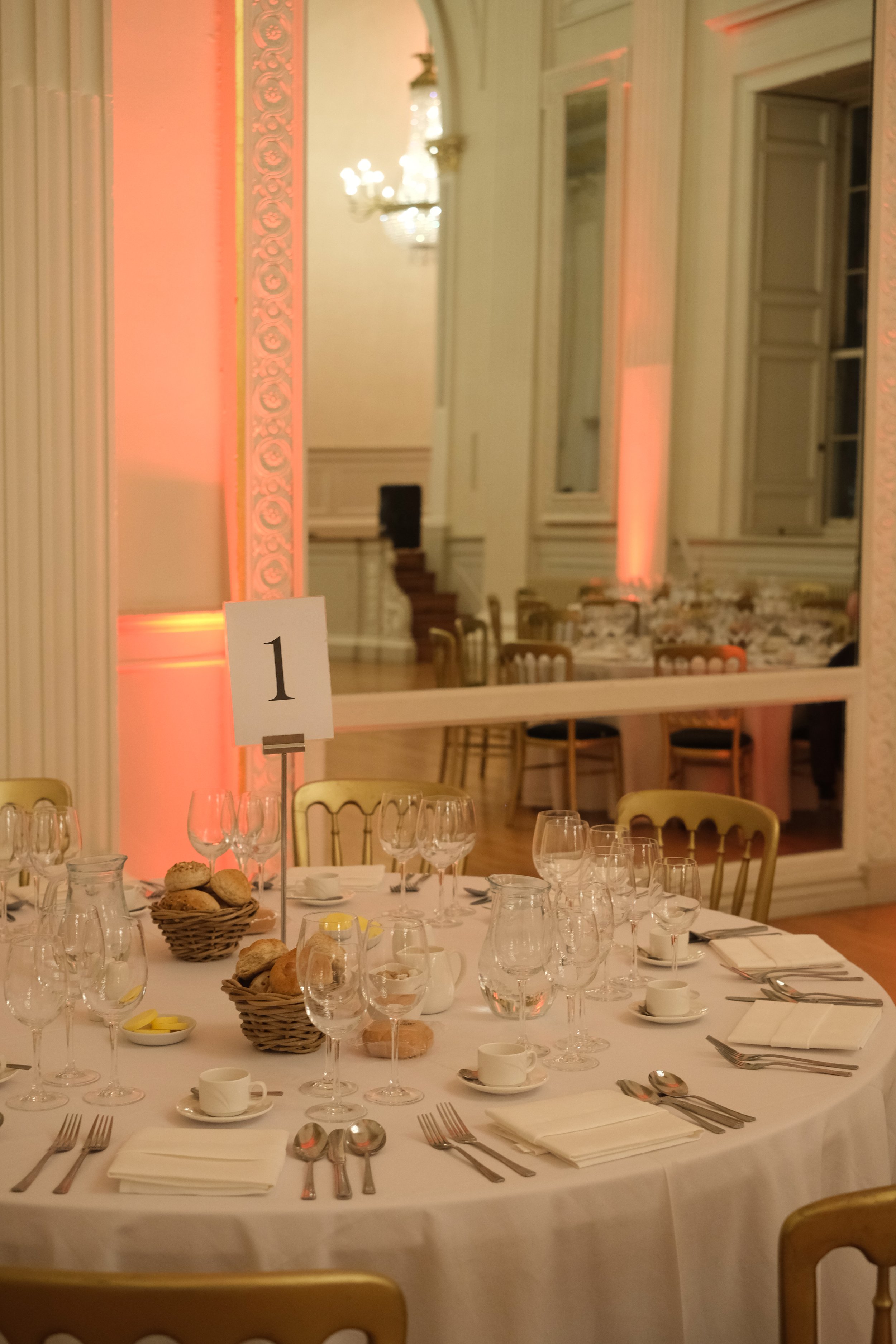 Elegant banquet table set with glassware, plates, silverware, cups, and napkins in a decorated room with warm lighting and a large mirror.