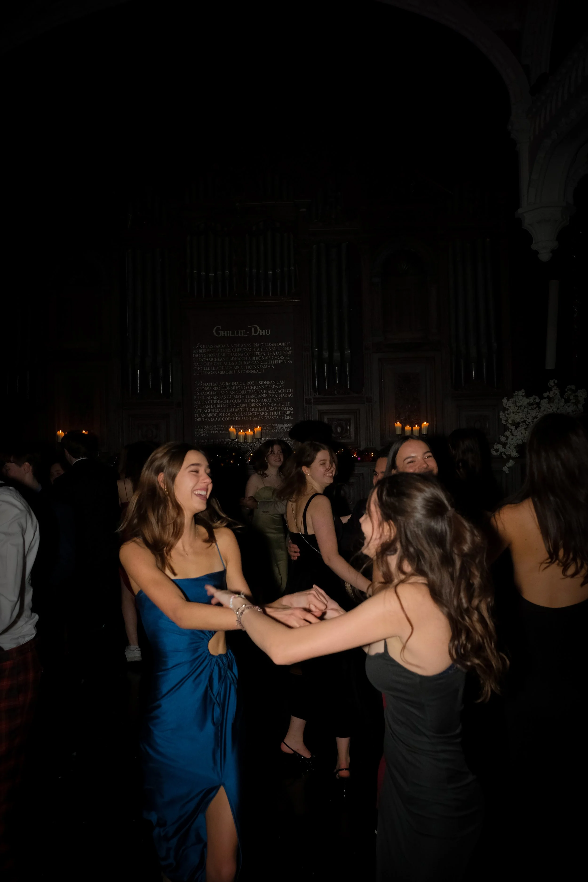 Young women dancing and smiling at a formal event in a dimly lit hall.