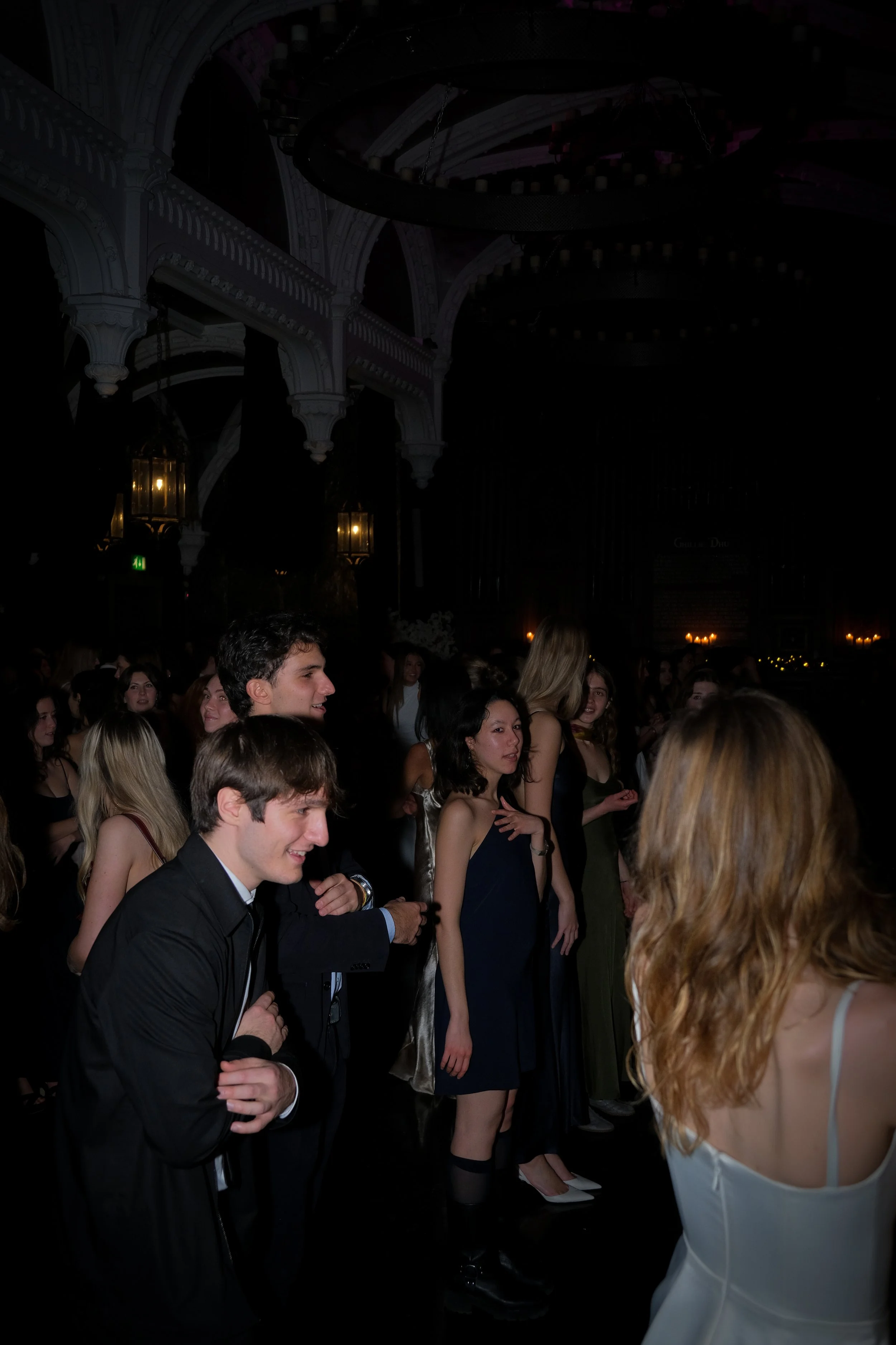 People dancing and socializing at a dark, elegant event or party, with ornate architectural details and warm lighting in the background.