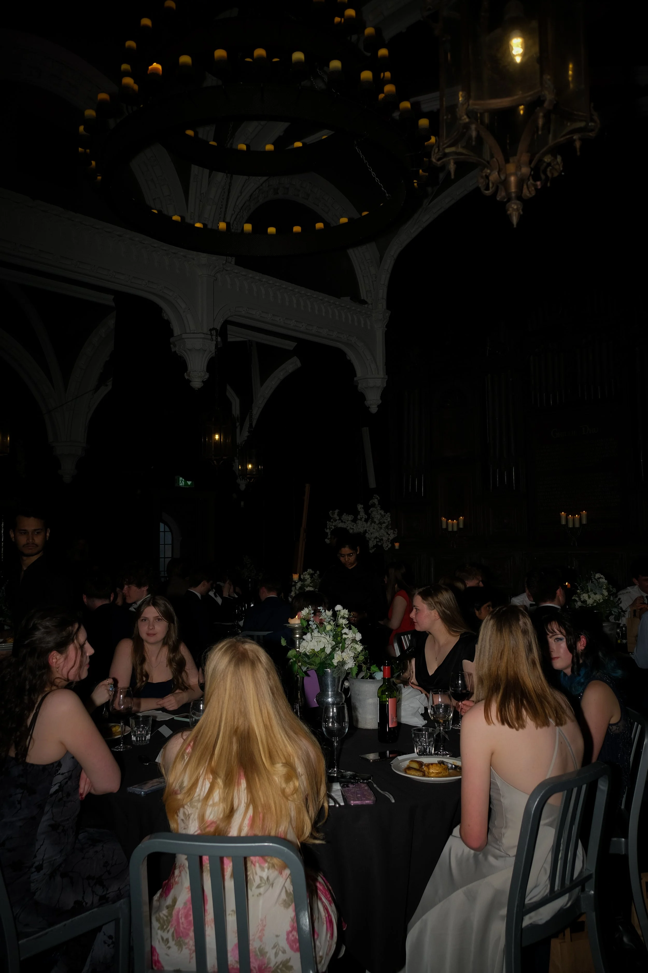 People sitting around a table at a formal event or dinner party, with floral centerpieces, wine bottles, and plates of food, in a dimly lit room with chandeliers.