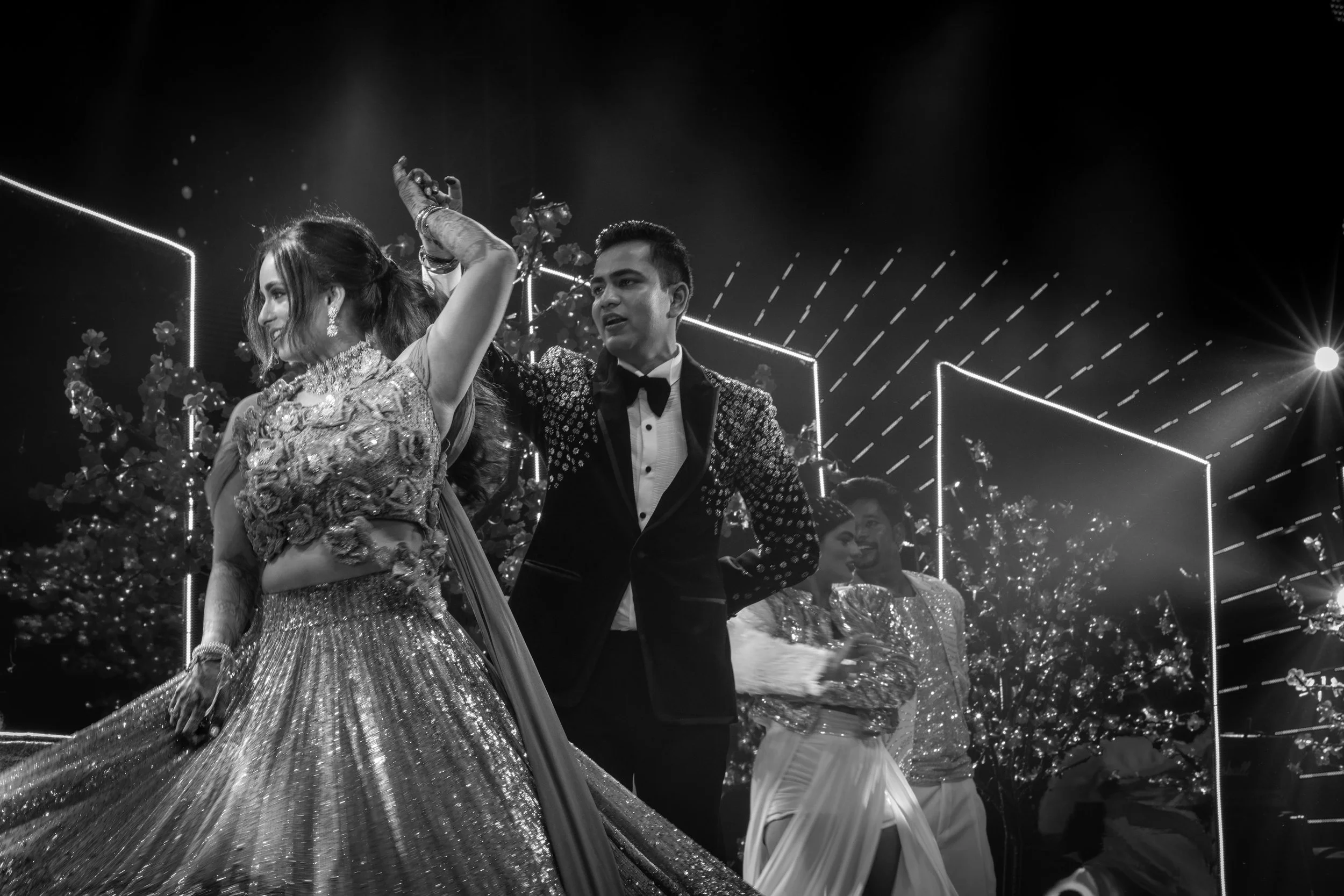 A black and white photo of a couple dancing at a celebration, with women in traditional Indian attire and men in tuxedos on a stage decorated with flowers and geometric light patterns.