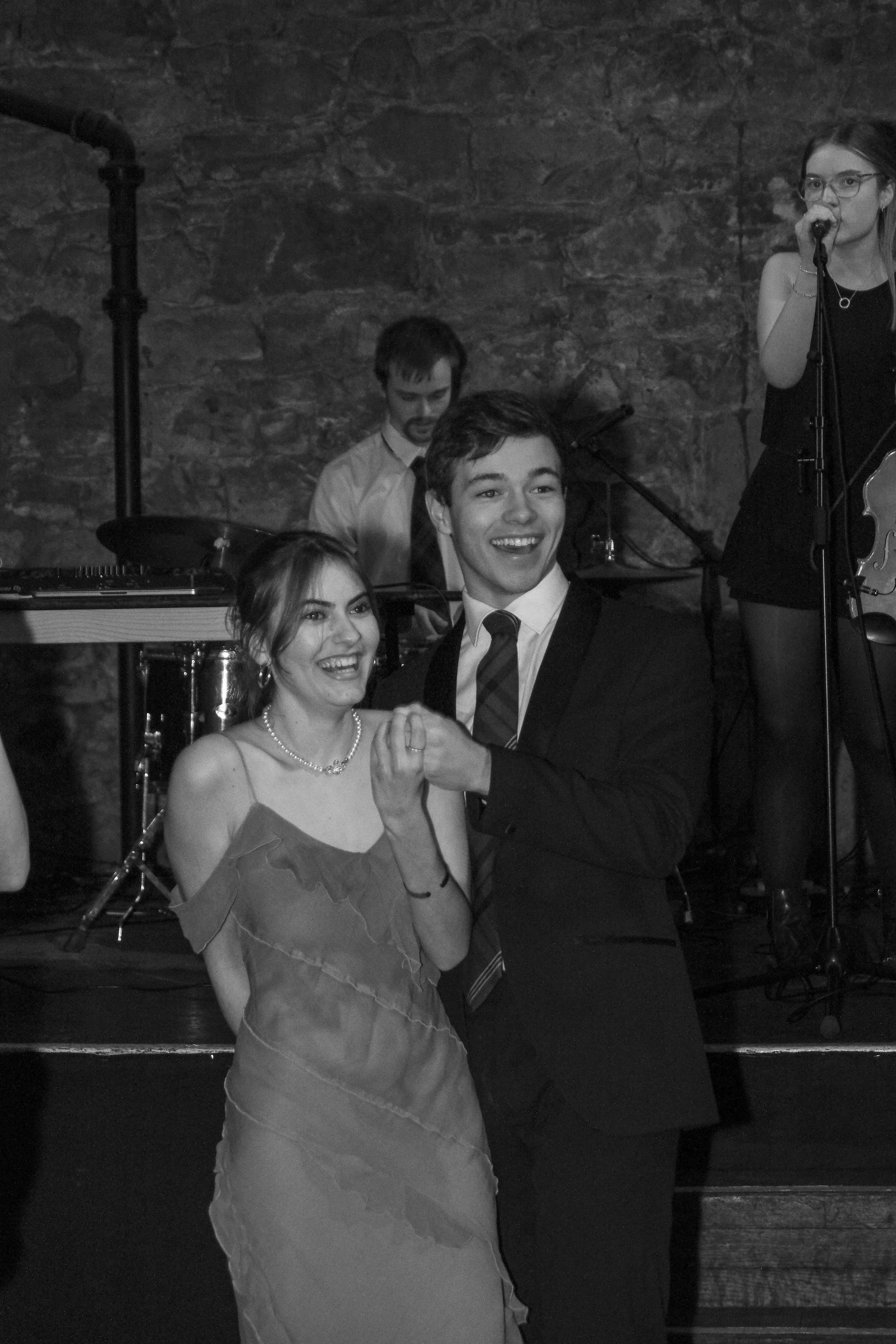 A black and white photo of a happy couple dancing together at a celebration, with a live band playing in the background.
