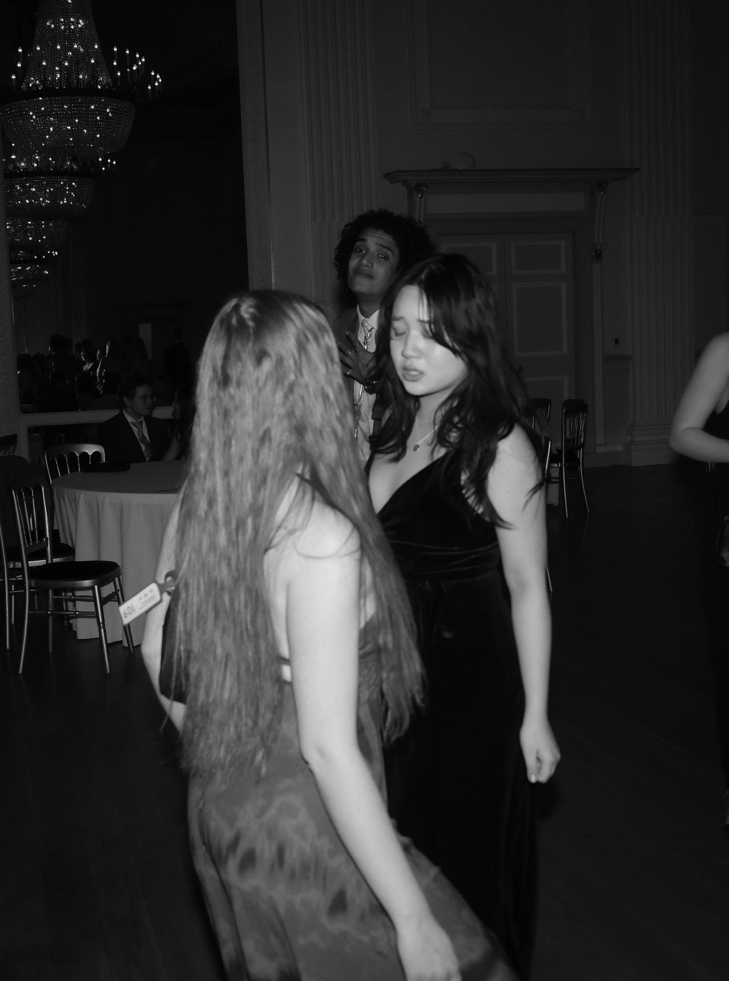 Two women dancing closely at a formal event, with a third woman in the background watching. The scene is in a ballroom with elegant decor, tables, chairs, and a chandelier.