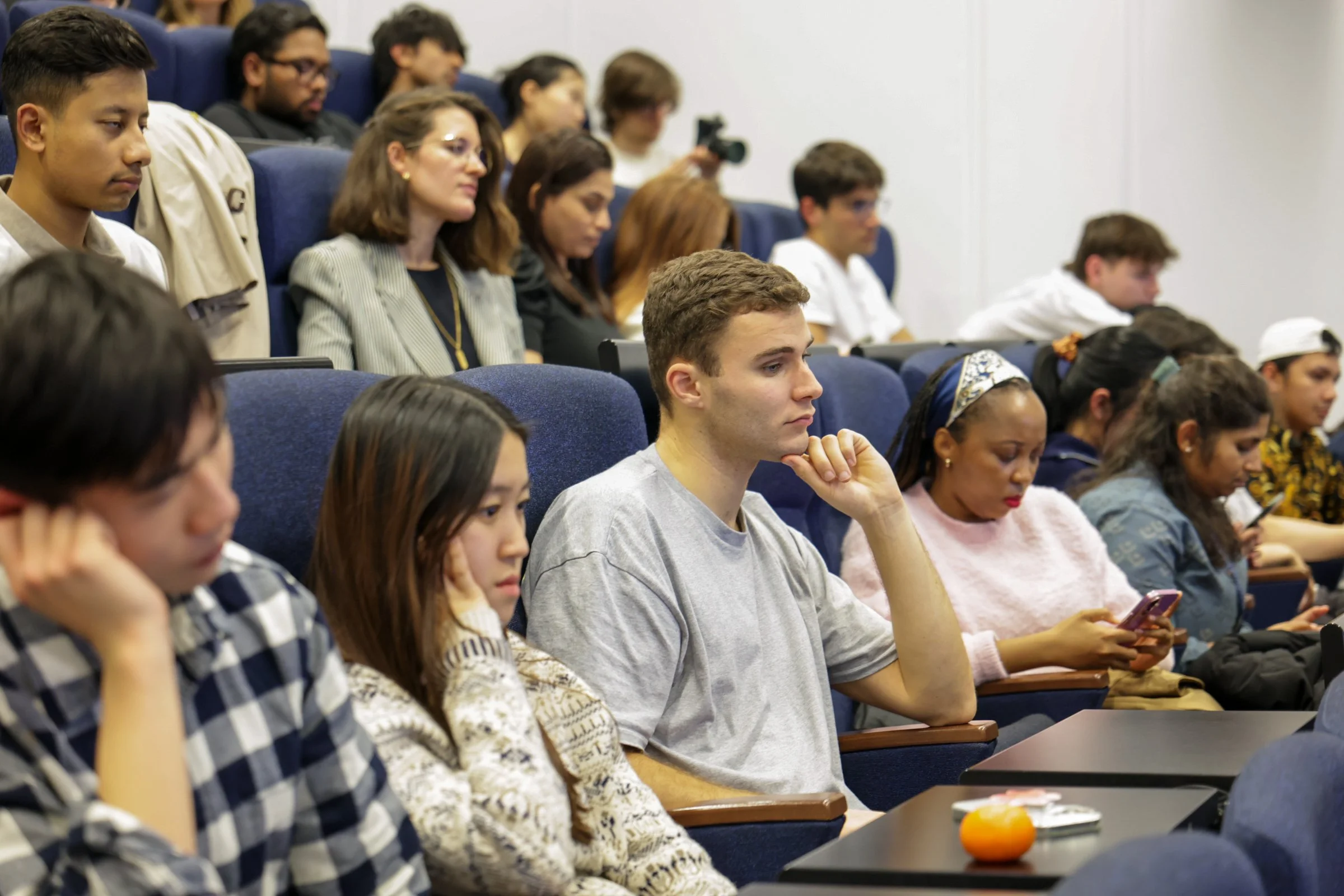 A diverse group of young adults attending a lecture or presentation in an auditorium, some focused on their phones, others listening attentively.