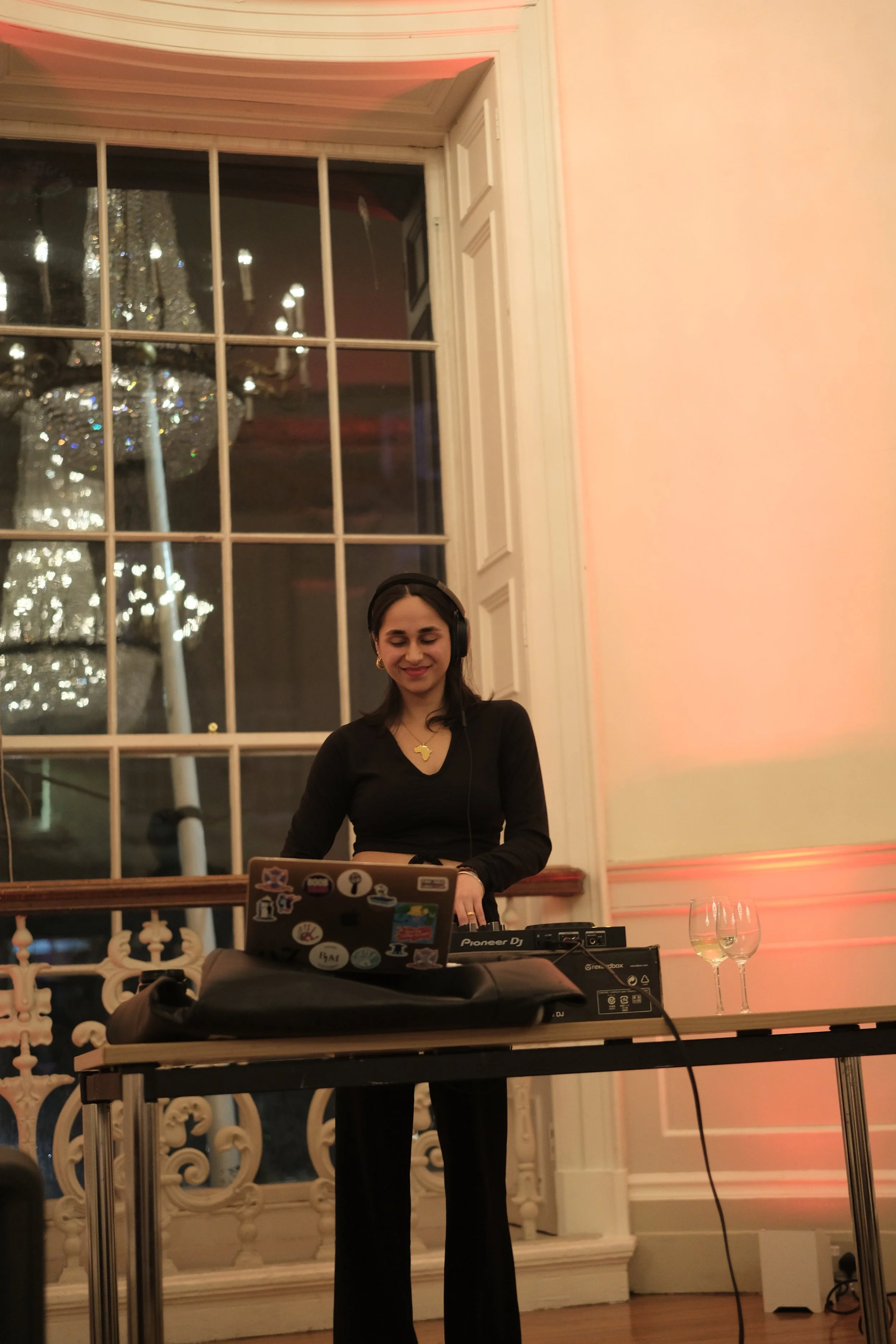 A woman wearing headphones and a black outfit standing at a DJ table with equipment, smiling. Two empty wine glasses on the table, large window and chandelier in the background.