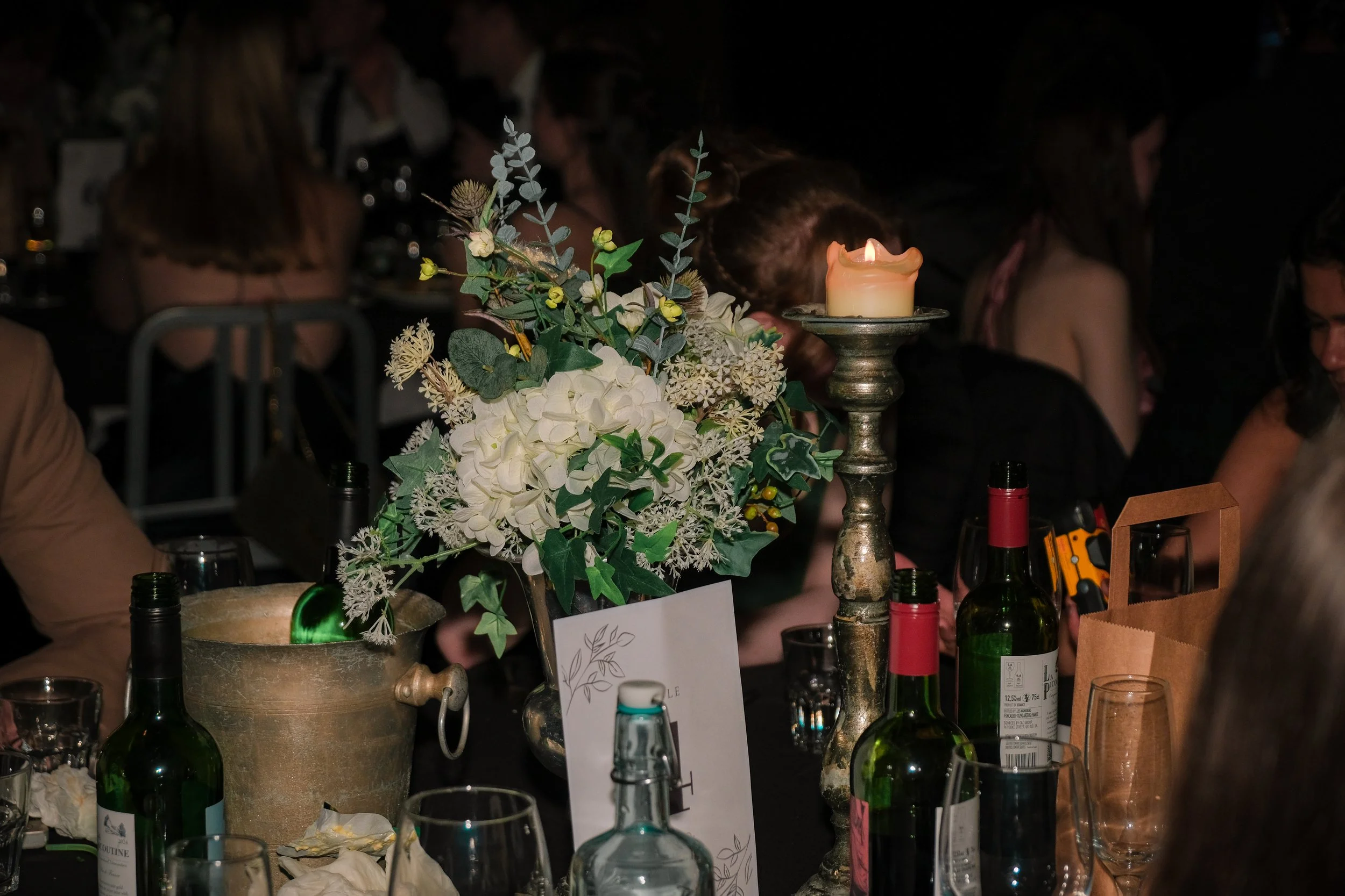 A candle on a stand, a floral centerpiece with white flowers and greenery, wine bottles, glasses, and a paper bag on a table at a dimly lit event with people in the background.