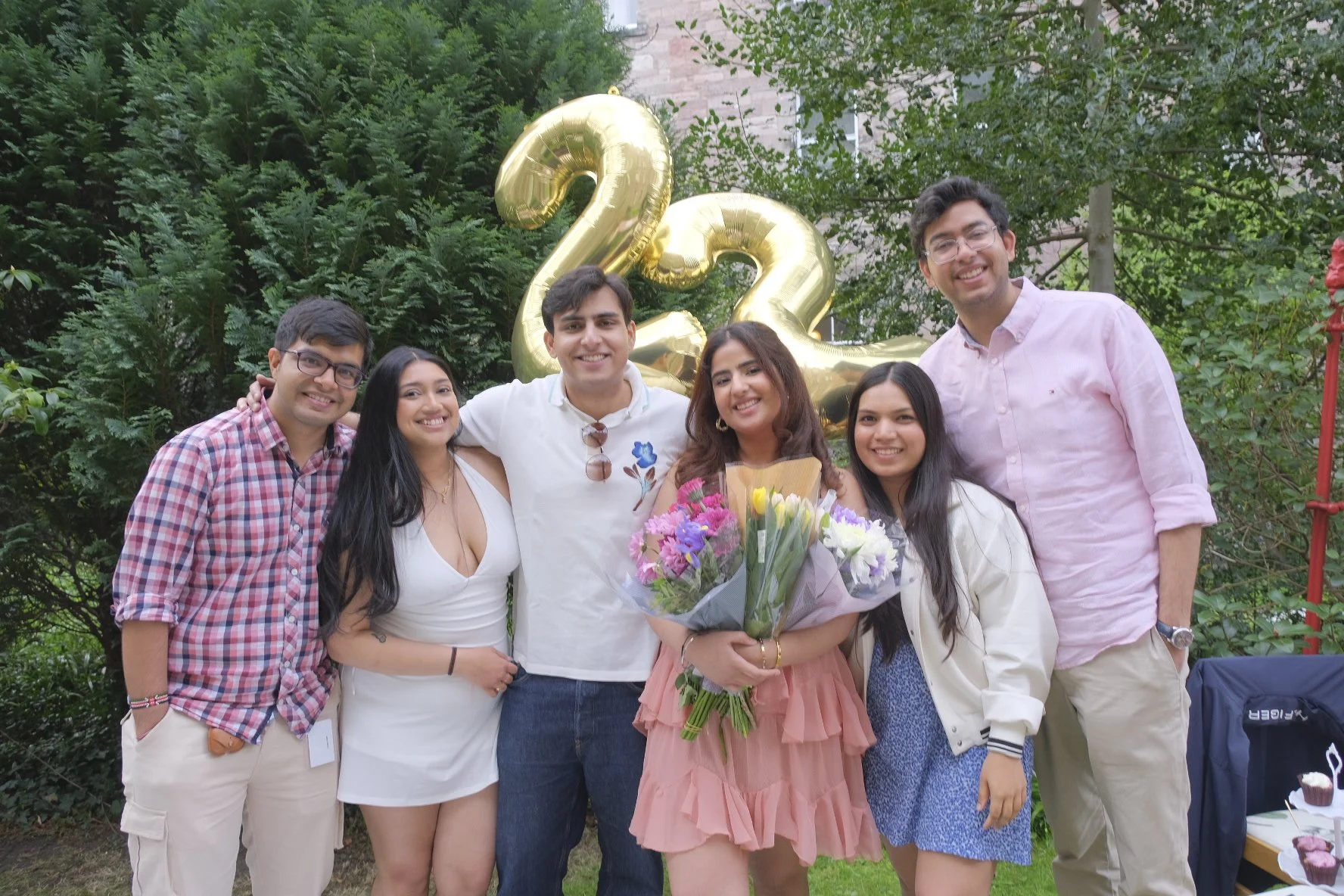 Group of friends celebrating with balloons and flowers, standing outdoors in front of large gold '22' balloons, amidst greenery.