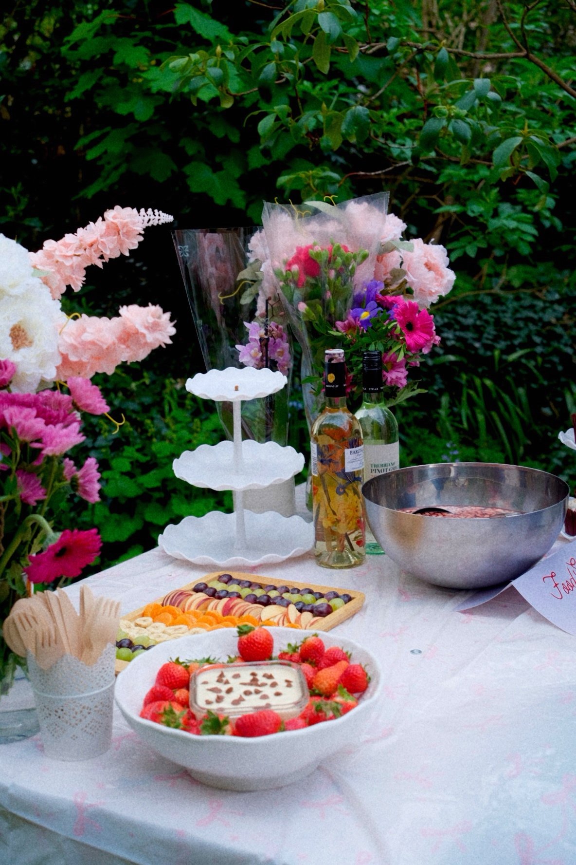 A table set outdoors with a variety of desserts, snacks, flowers, and beverages, including strawberries, a layered cake, chocolates, a bowl, and bottles of wine or liquor.