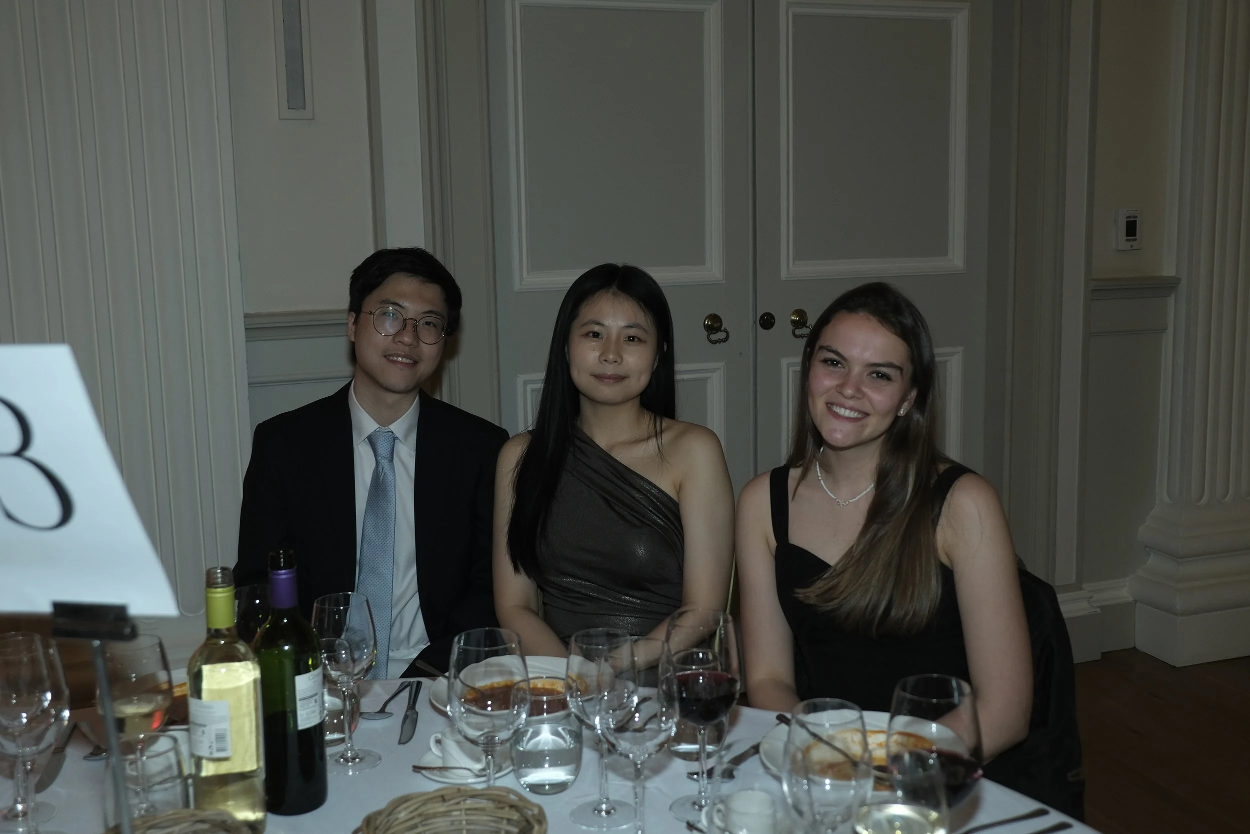 Three young adults seated at a table in a formal setting, smiling, with wine glasses and bottles on the table, white paneled walls in the background.