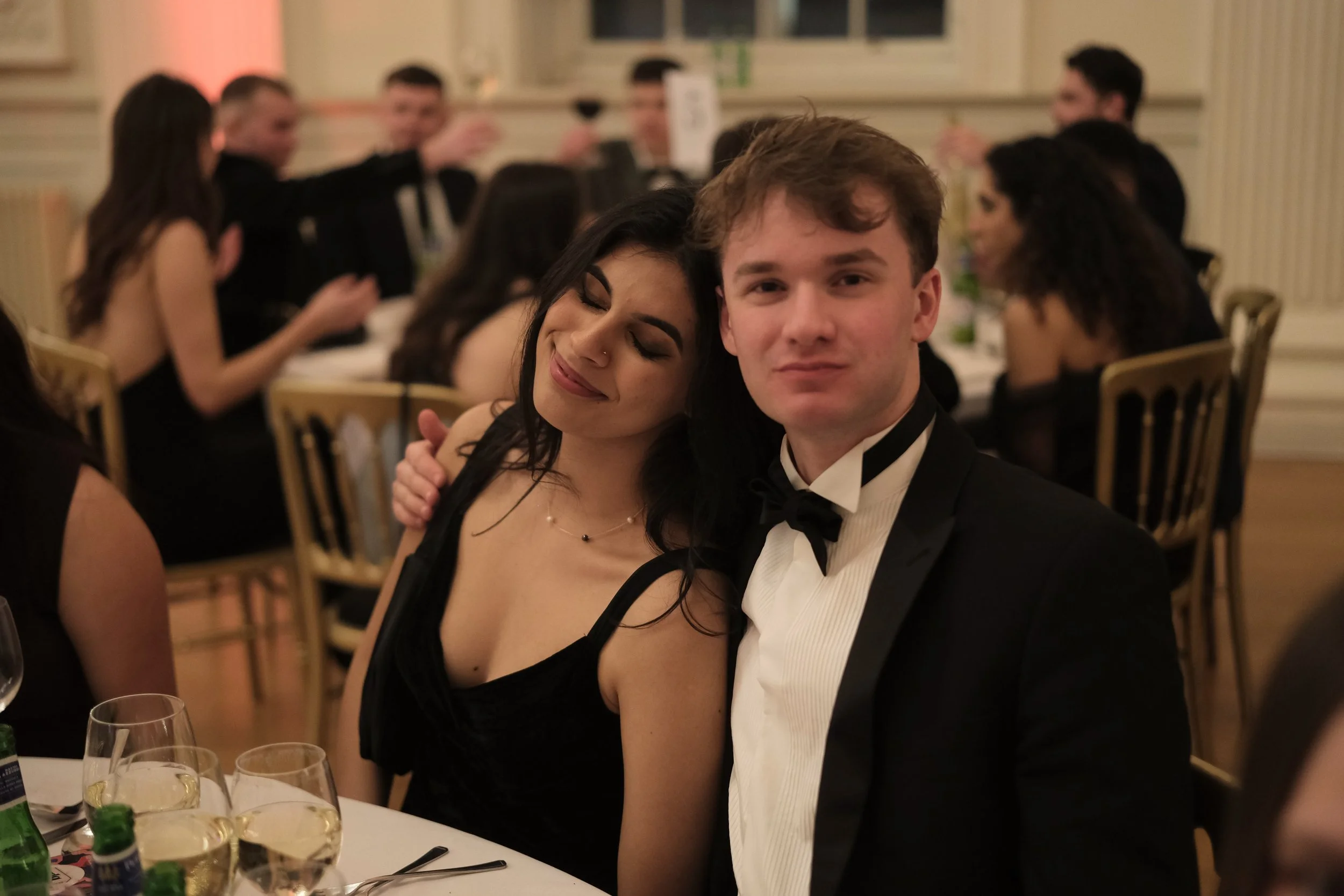 Two young adults, a woman with dark hair and a man with light hair, dressed in formal attire, sitting at a table during an elegant event or banquet. The woman has her eyes closed and is smiling gently, with her head resting on the man's shoulder. The