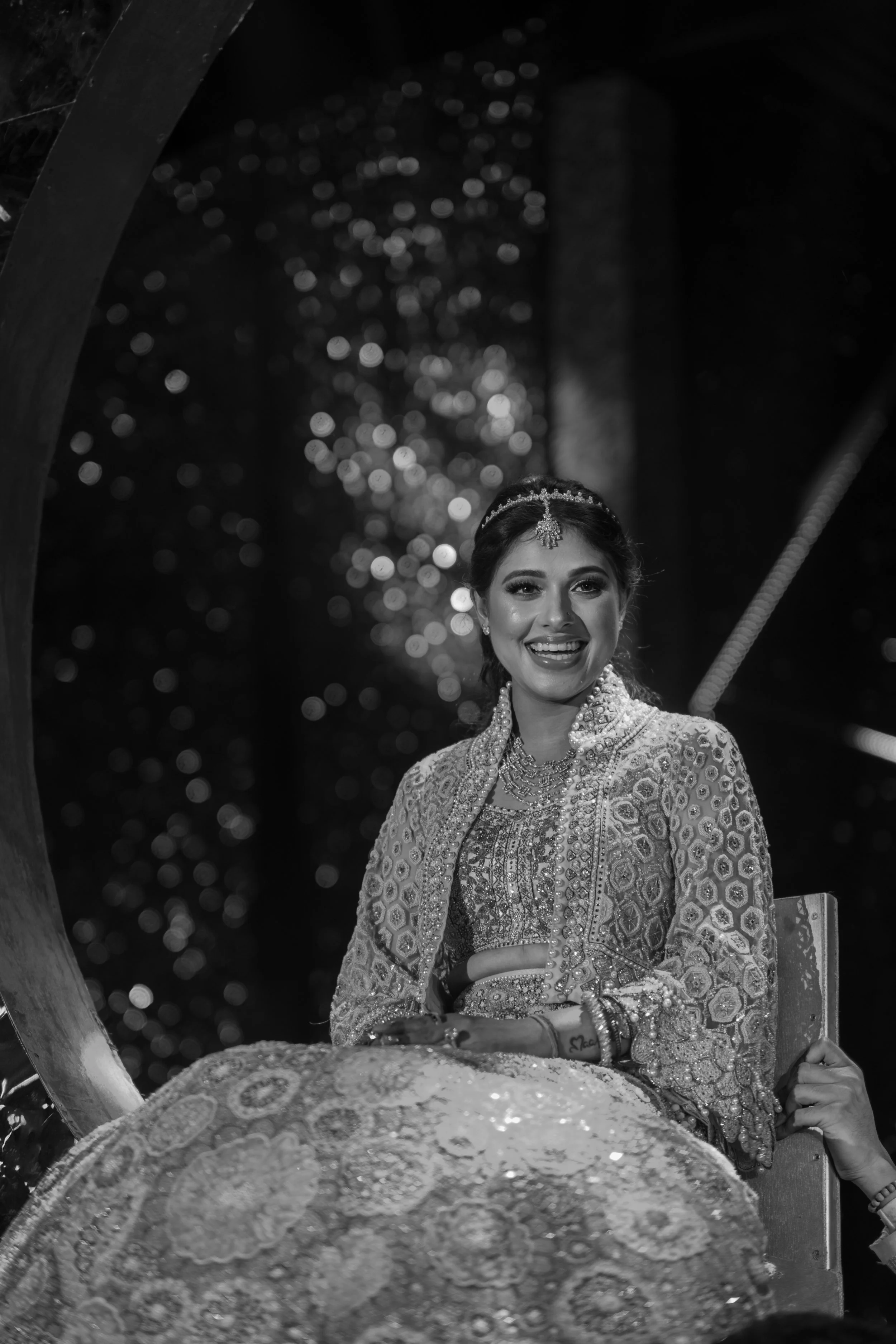 A woman dressed in ornate traditional attire, sitting on a decorated chair, smiling, with intricate jewelry and a tiara, in a nighttime setting with bokeh lights in the background.