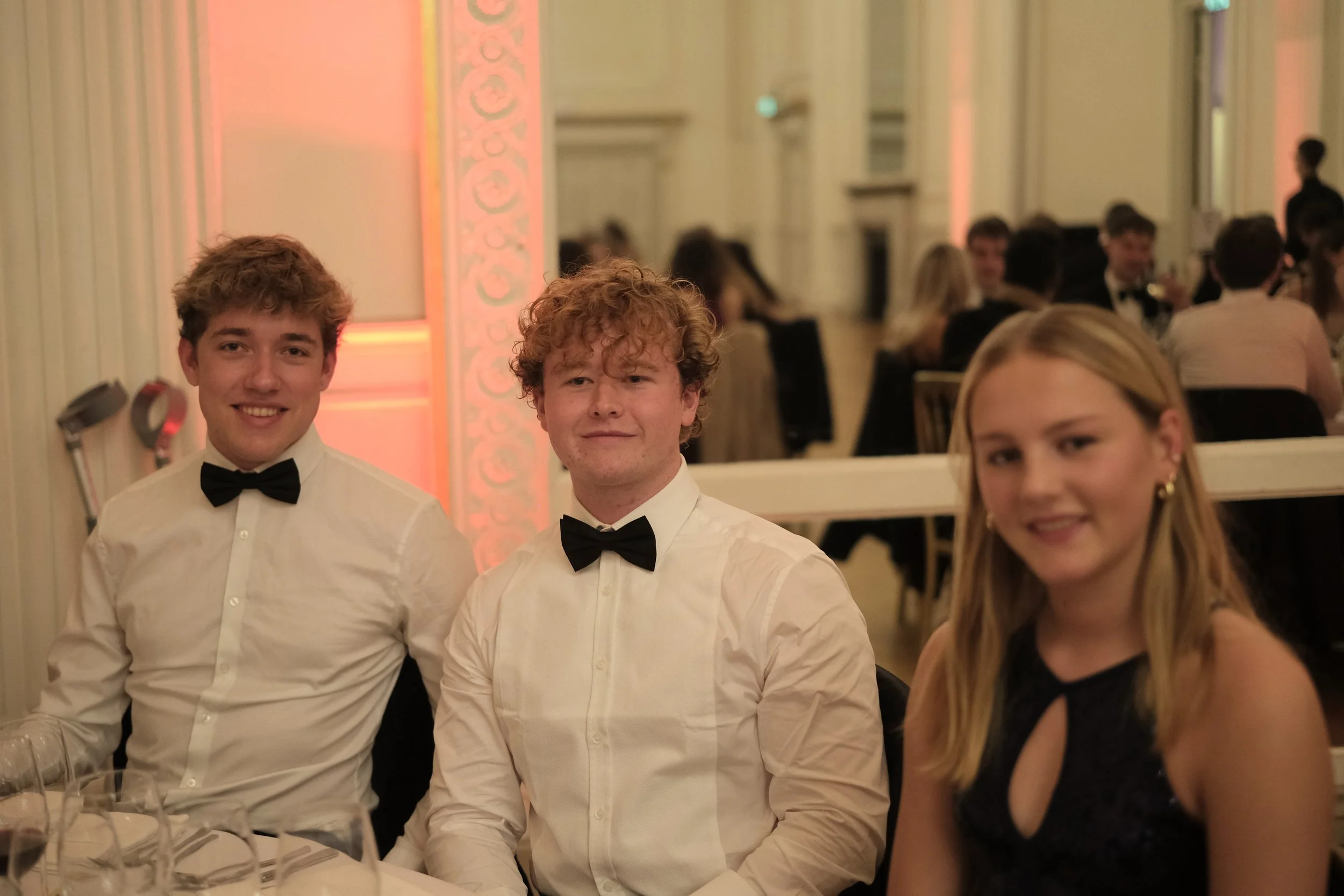 Three young adults in formal attire sitting at a dinner table during a celebration or event, with a crowd of people in the background.