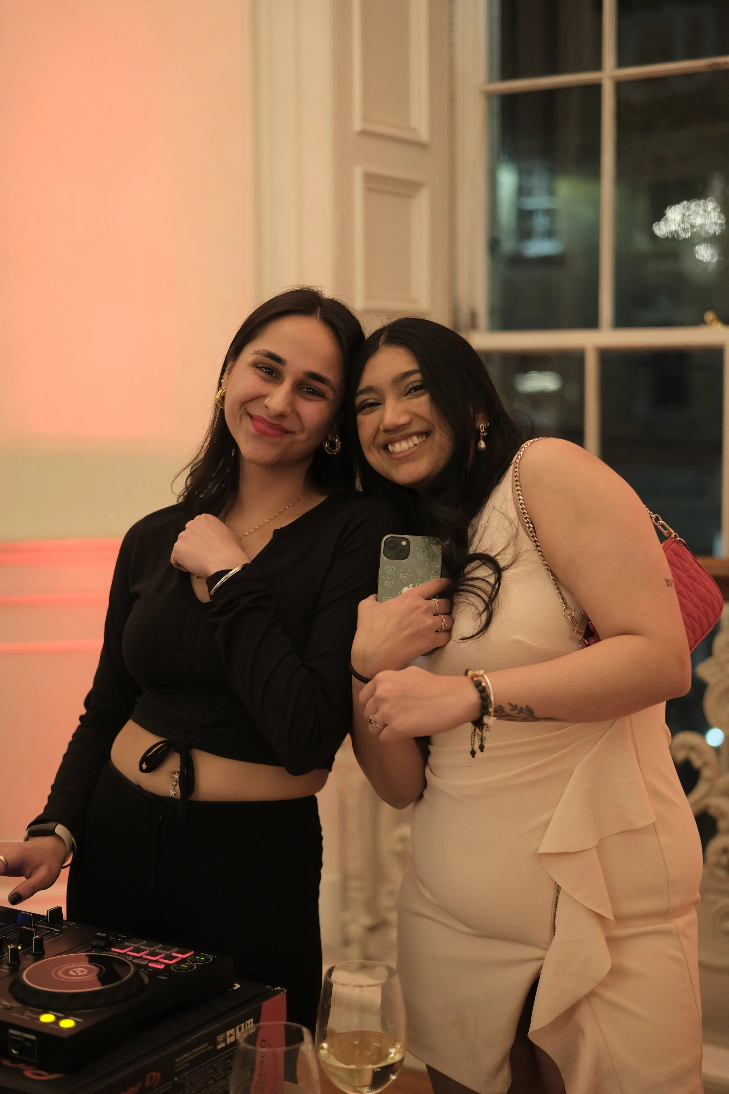Two women smiling and taking a mirror selfie at a party or celebration, with one holding a smartphone and a DJ equipment on the table in front of them.