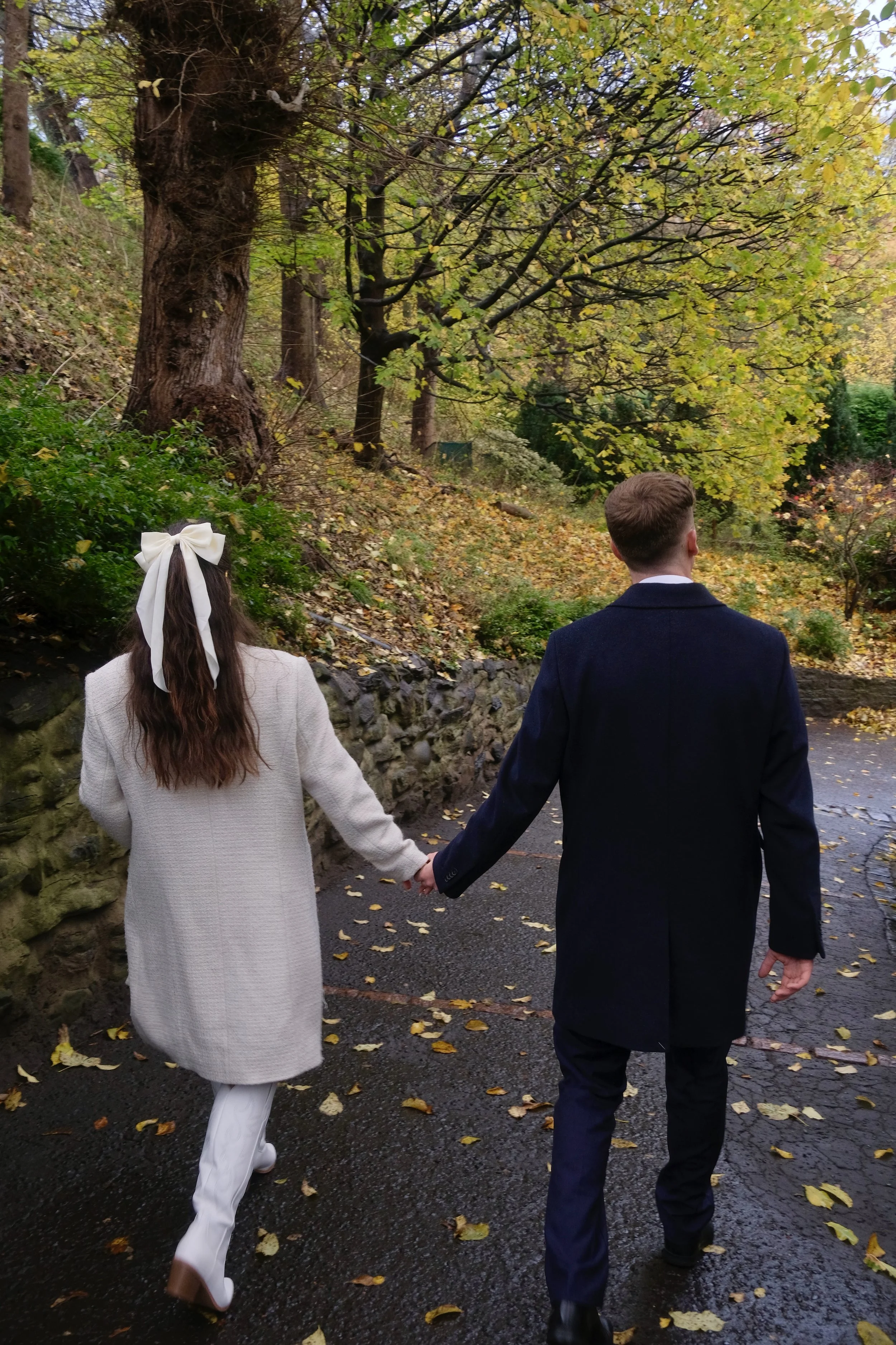 A couple walking hand in hand through a park with autumn leaves on the ground, trees with yellow and green leaves, and a stone wall along the path.