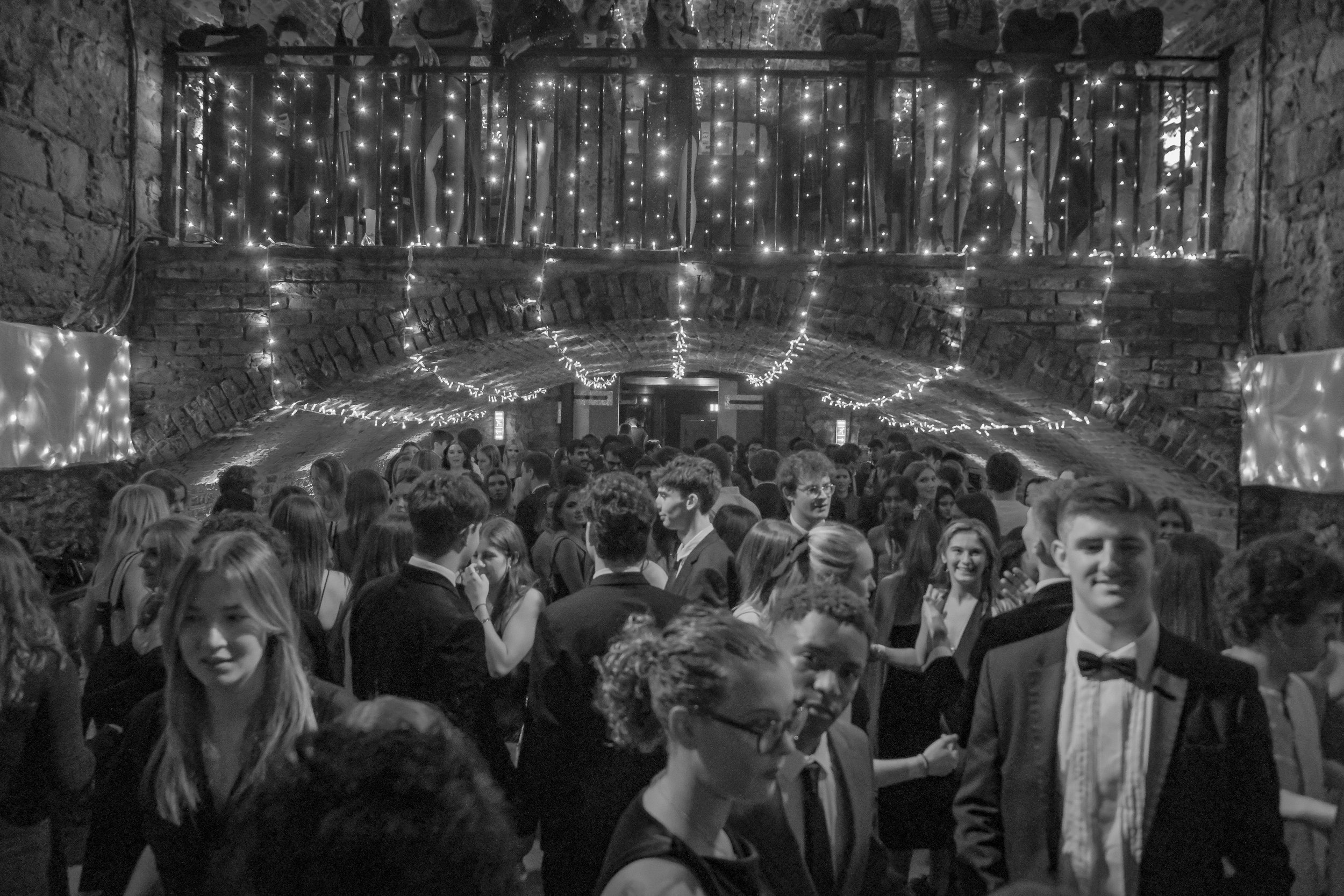 A black and white photo of a crowded ballroom or event space with many young people dressed formally in tuxedos and dresses, dancing and socializing beneath string lights and a balcony with additional attendees overlooking the scene.
