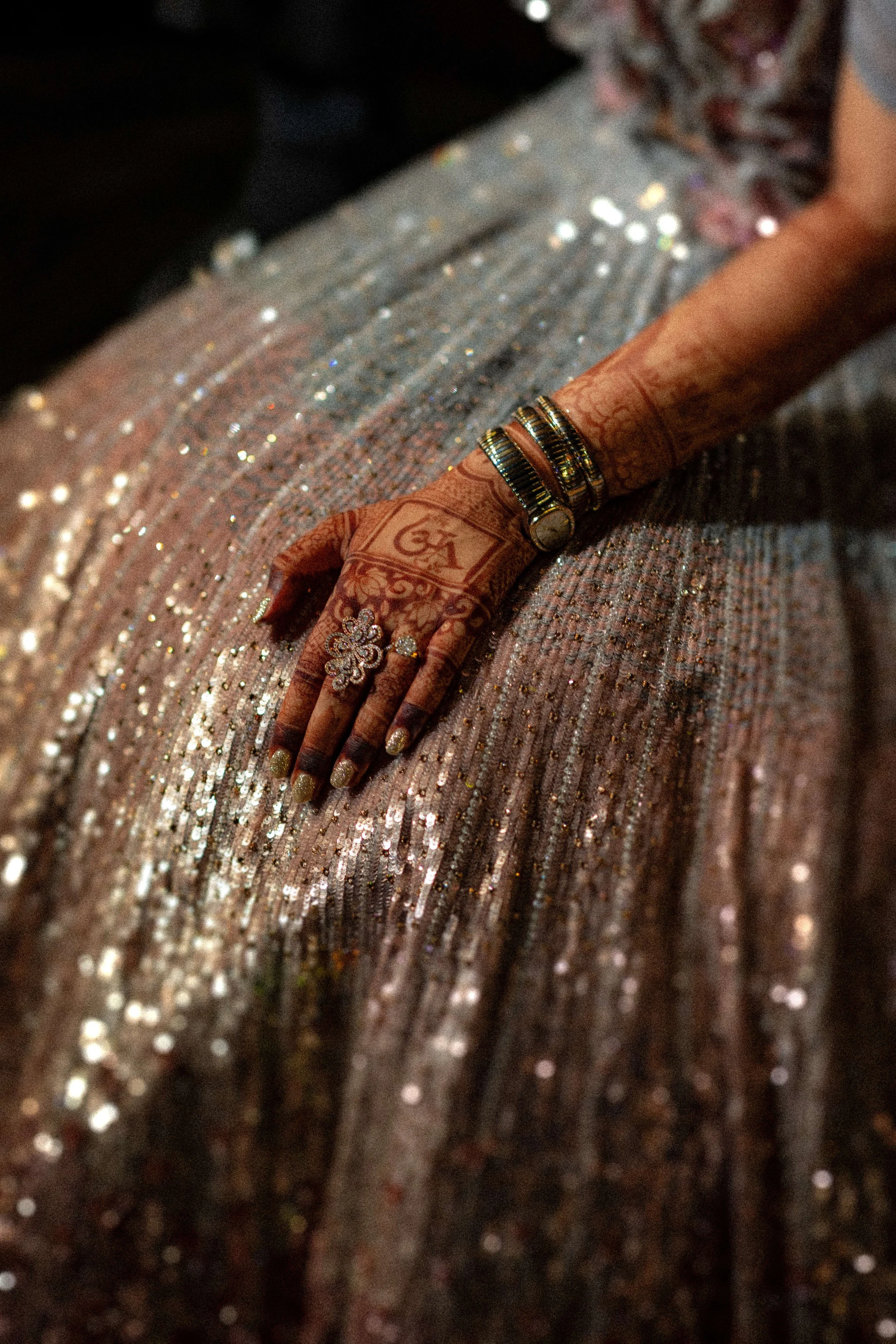Close-up of a woman's hand adorned with jewelry, resting on a shiny, gold sequin dress. The hand features henna designs and is decorated with rings and bangles.