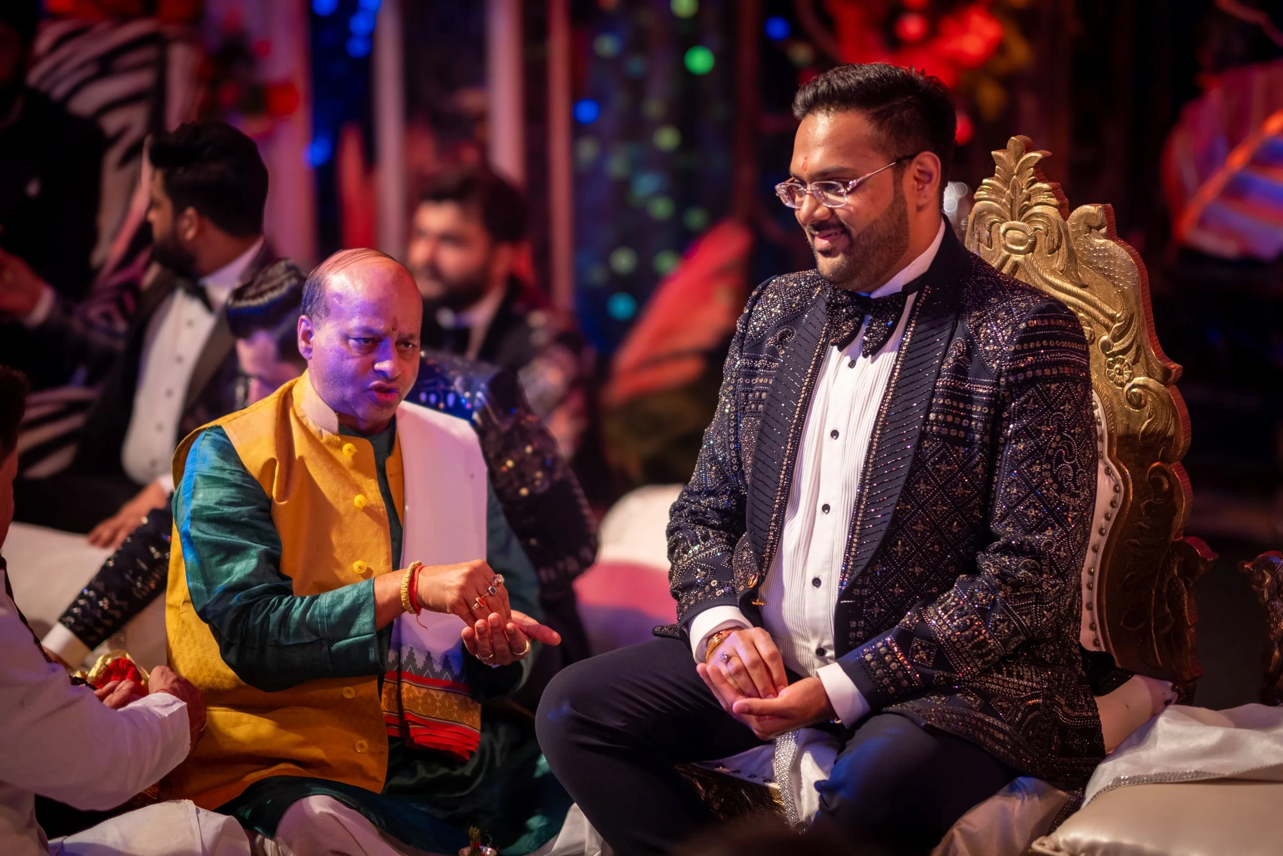 A groom in a black and white tuxedo sitting on a gold ornate throne at a wedding, smiling and clasping his hands, while an older man in traditional Indian attire talks to him during the celebration.