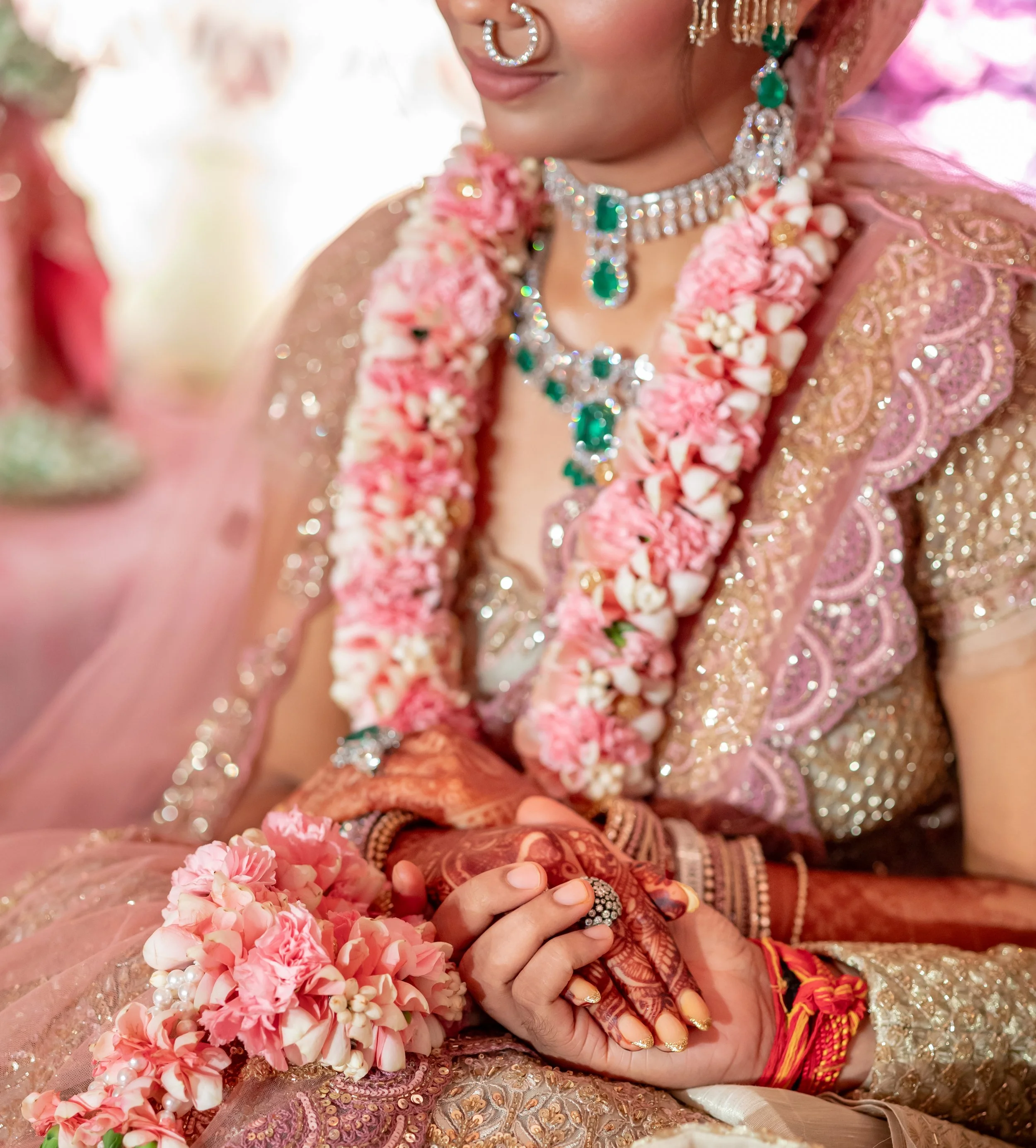 A woman wearing colorful traditional Indian jewelry, including a nose ring, necklace, earrings, and bangles, is dressed in a pink and gold embroidered outfit. She has flowers around her neck and wrist, and henna on her hand. The photo focuses on her upper body and jewelry.
